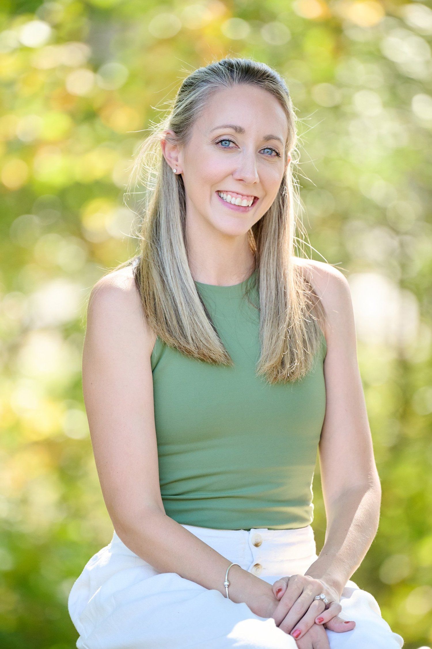 A person wearing a sleeveless green top and white pants sits outdoors with a natural blurred background.