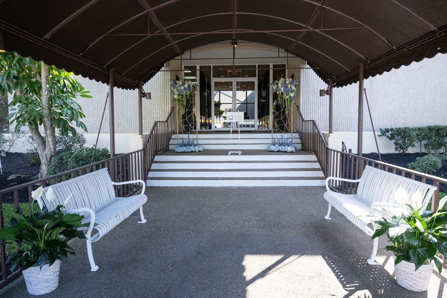 A covered outdoor walkway with white railings and potted plants leads to a building entrance.
