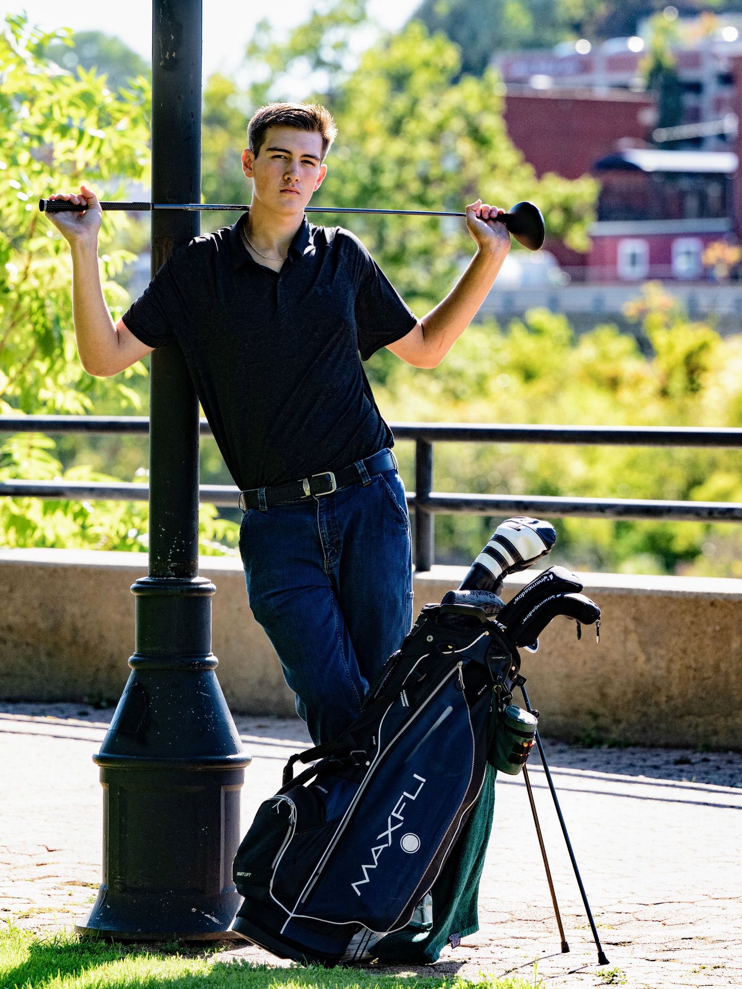 A golfer in dark attire sits beside a golf bag on a brick wall overlooking a scenic landscape on a sunny day.
