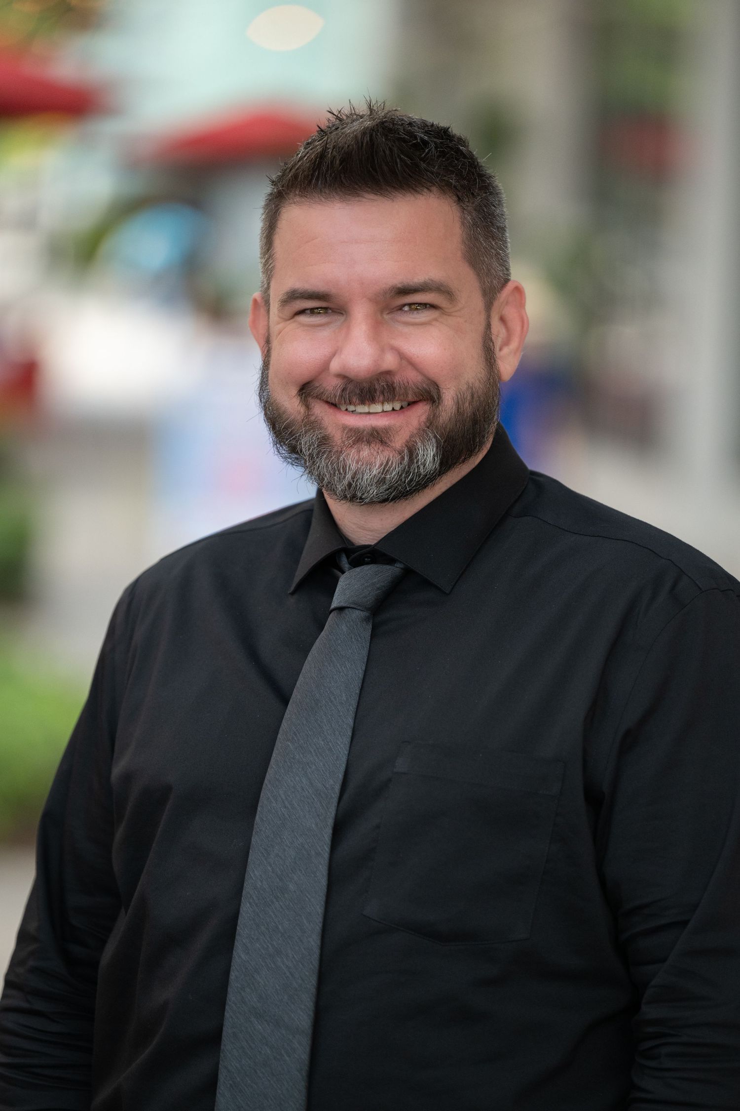 Smiling man with beard wearing black shirt and gray tie poses outdoors in professional headshot photo.