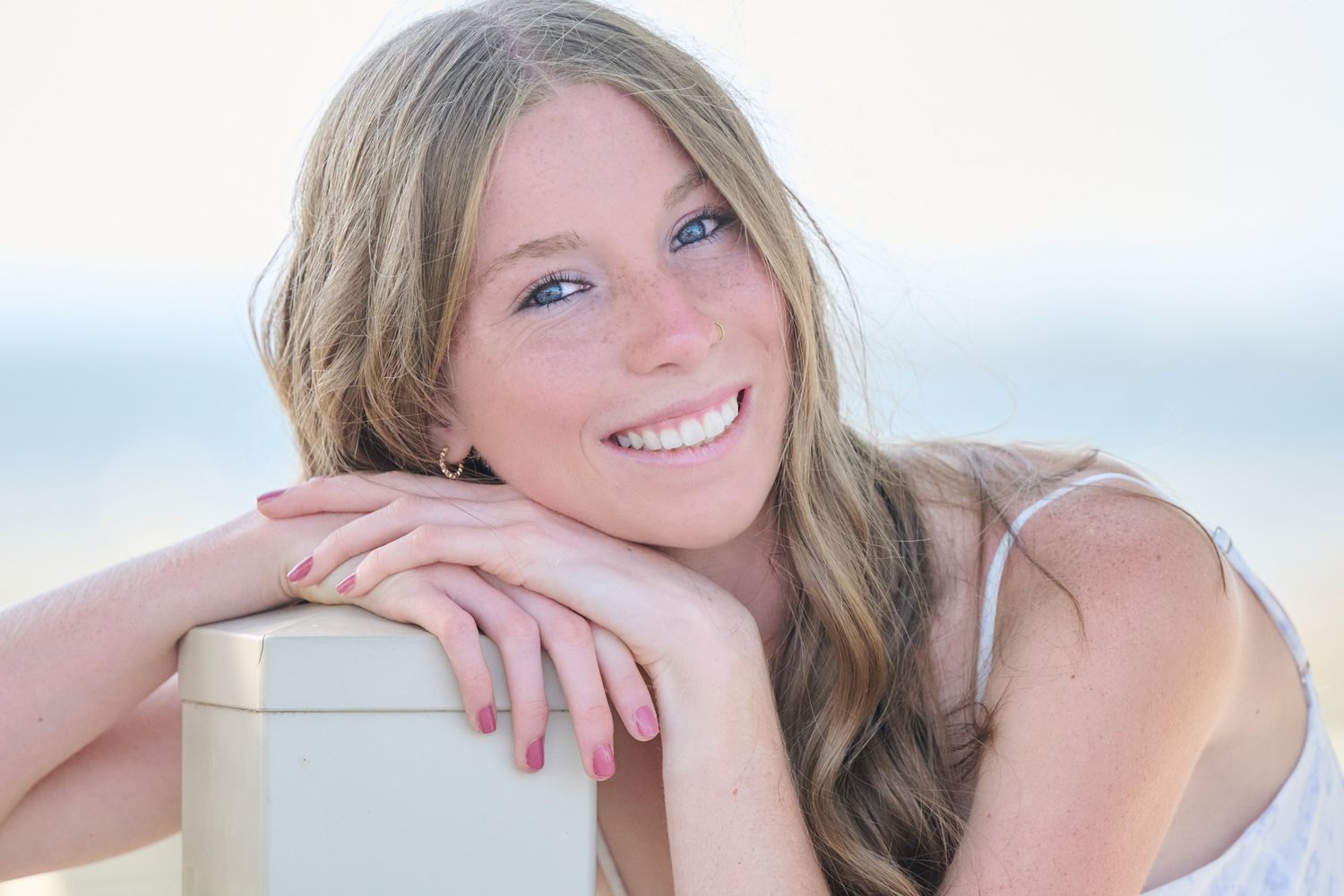 A person with blonde hair poses outdoors in bright natural light wearing a white top.