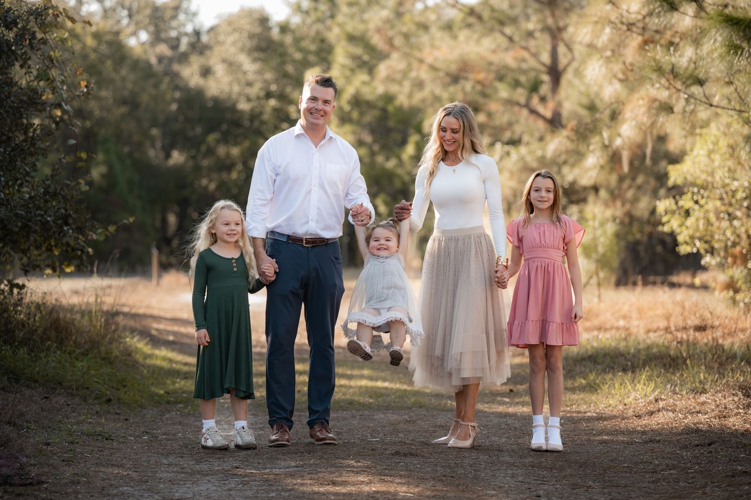A group of children in formal attire walk together on a dirt path surrounded by trees during golden hour lighting.
