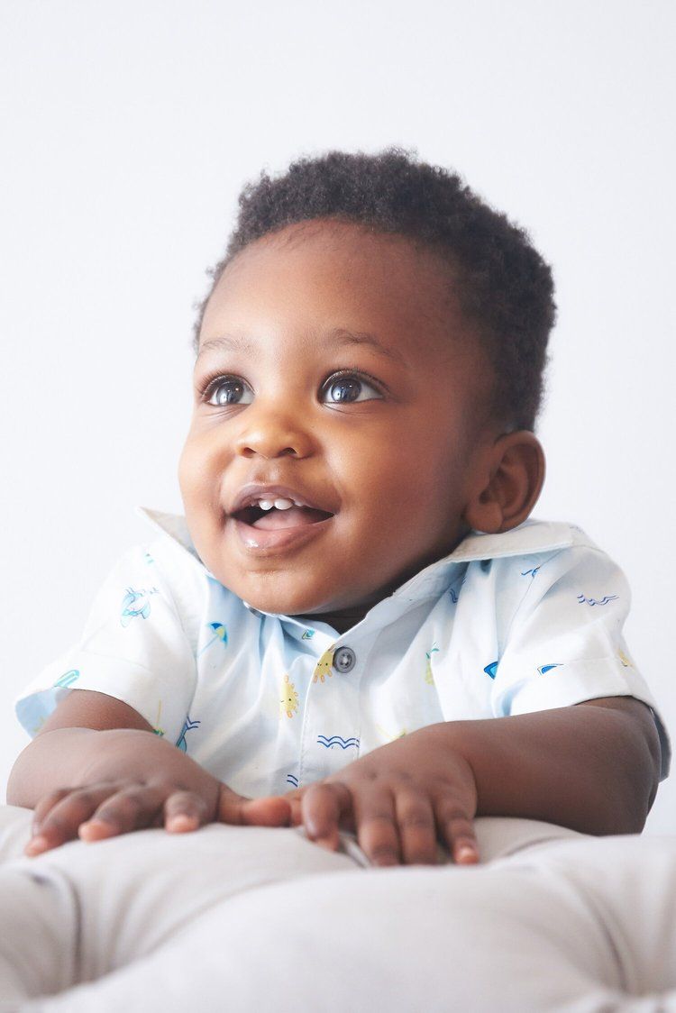 Adorable baby wearing white patterned shirt smiles brightly while lying on light-colored surface.