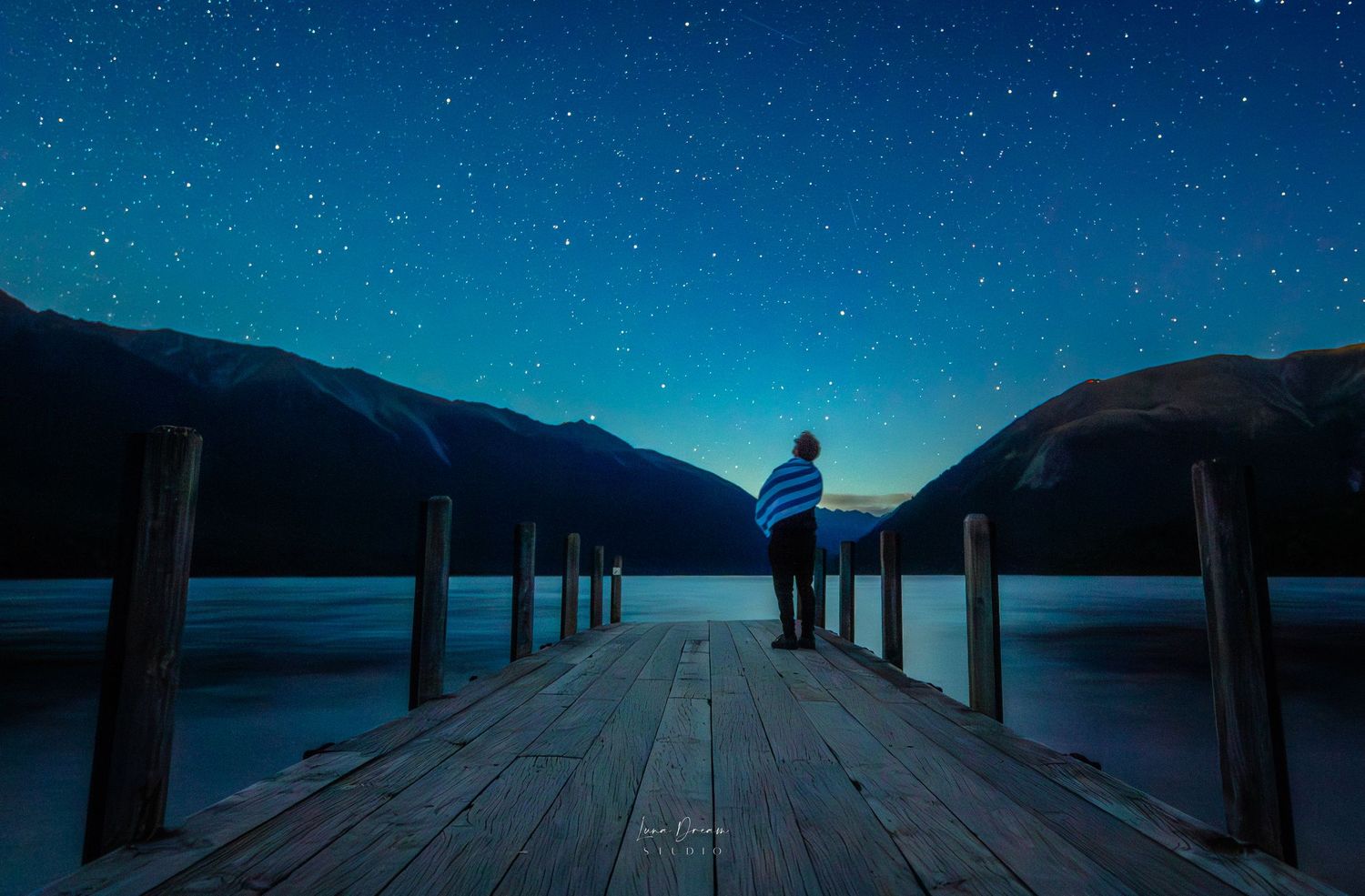 Nelson Lake New Zealand. A lone figure standing on a quiet pier under a deep starry sky, capturing peaceful nightscape, and serene lakeside
