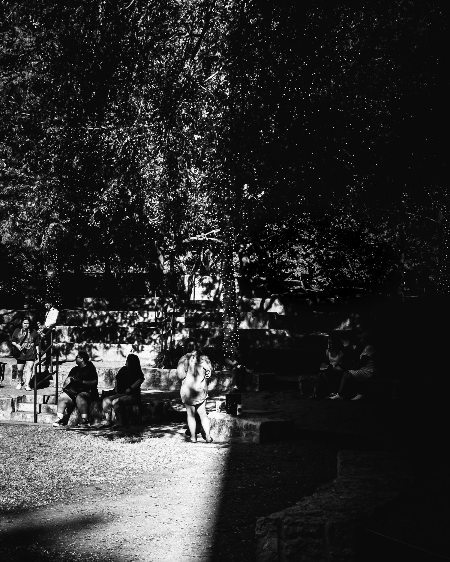 Black and white photograph of people gathering in a shaded park area.