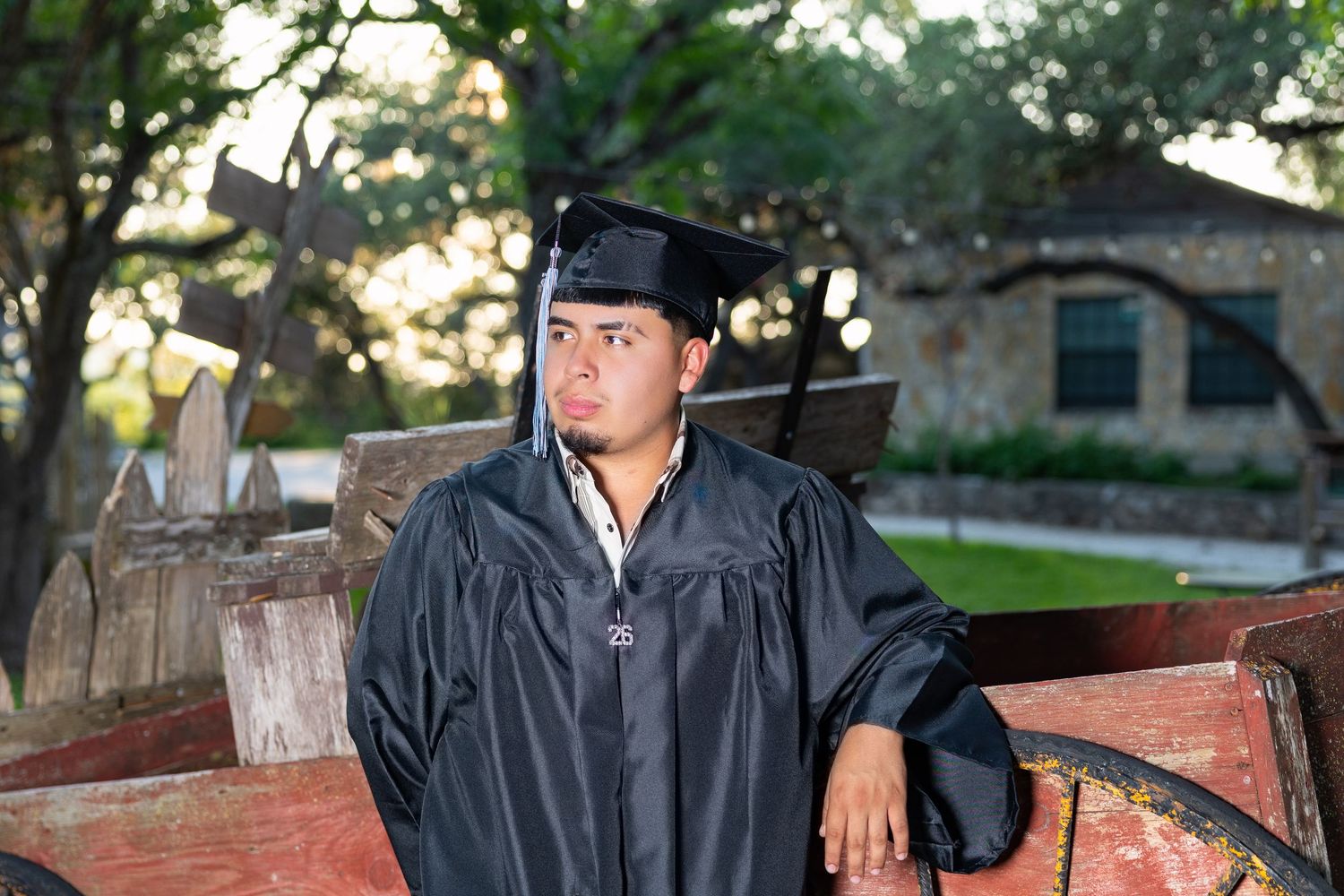 Young man in black graduation cap and gown posing outdoors near rustic wooden cart and trees.