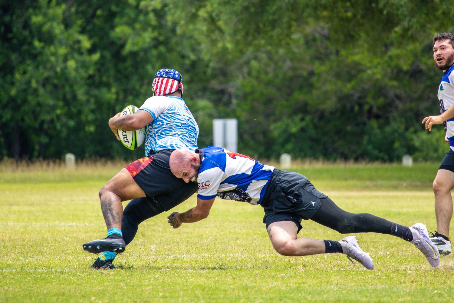 A rugby player in a patriotic hat gets tackled low by an opponent in blue and white during an outdoor match.