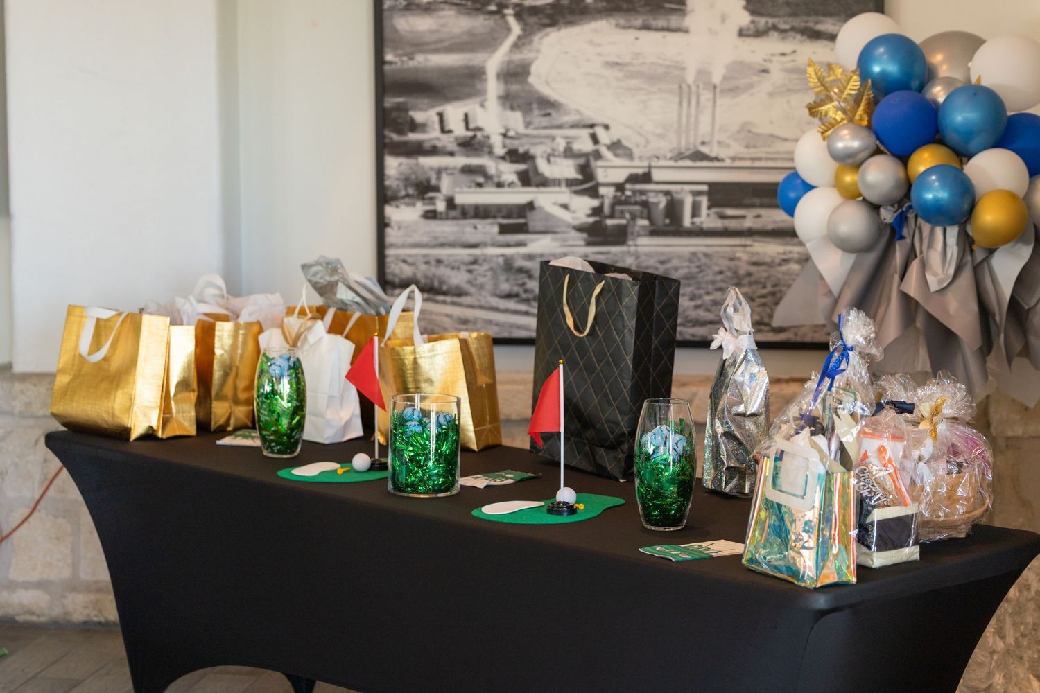 A decorated table with gold gift bags, golf-themed decorations, and blue and gold balloon arrangement at an event.