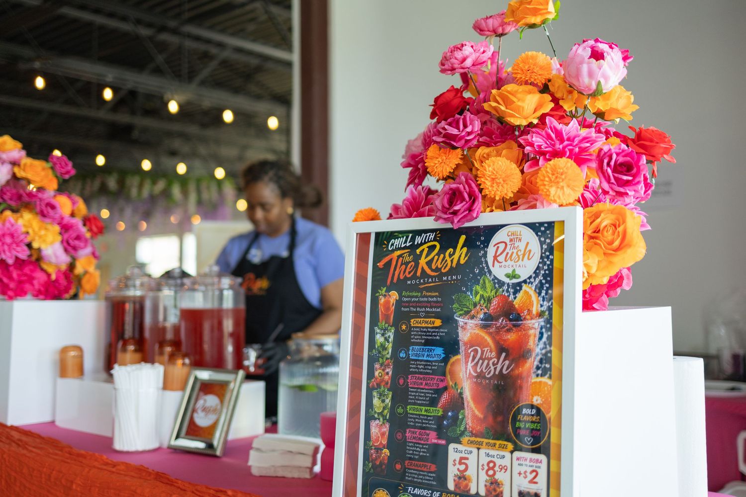 Decorative floral display and menu sign for The Rosh drink vendor at an indoor market event.