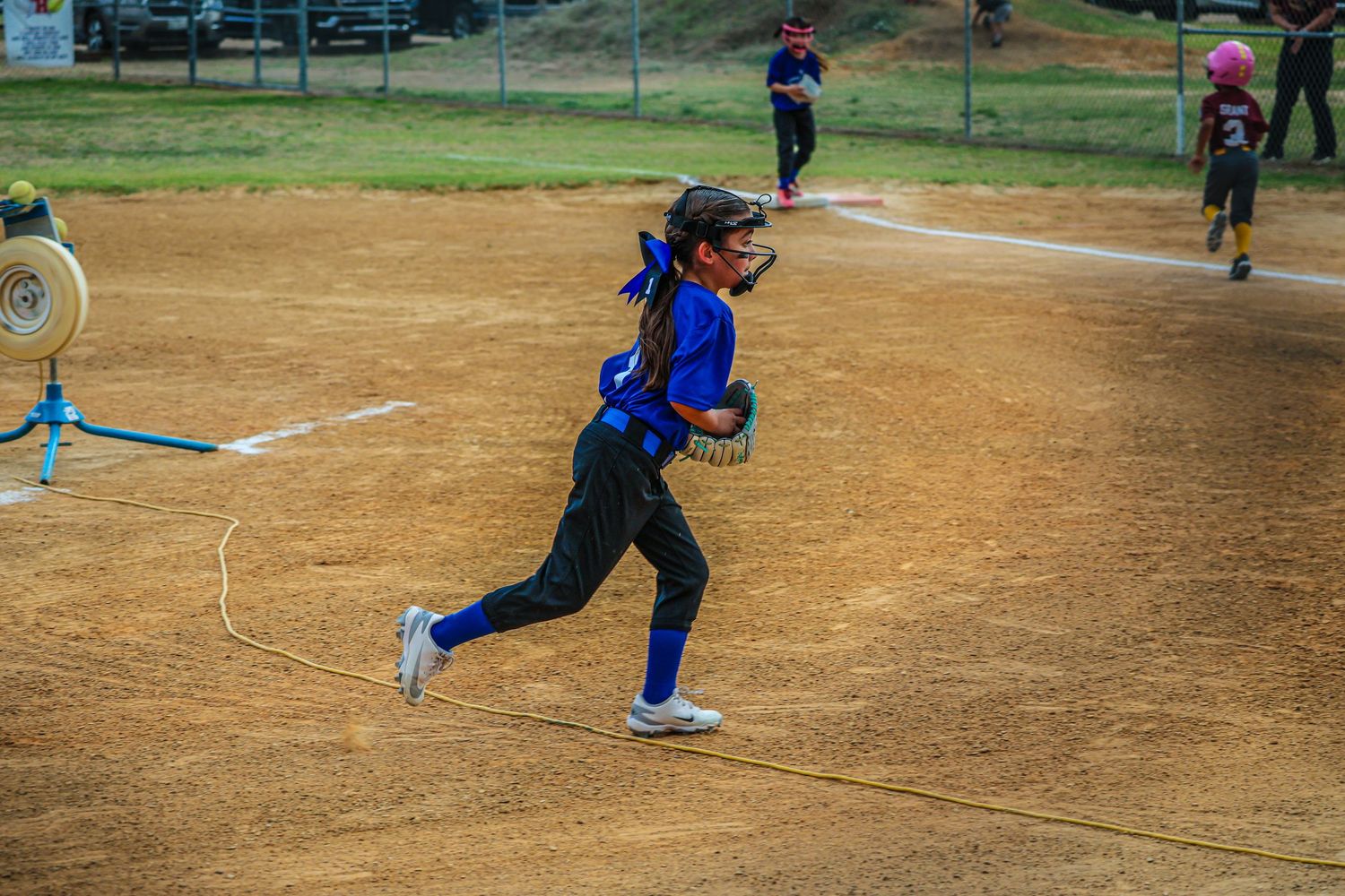 Young girl in blue uniform running bases during a youth softball game on a dirt field.