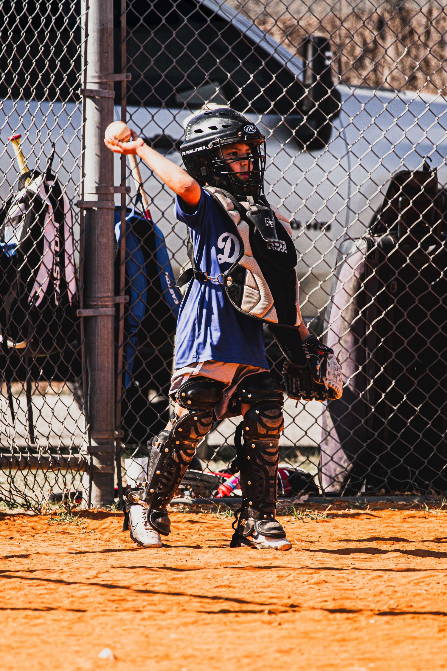 Youth softball catcher in full gear including helmet and chest protector stands ready on dirt field near chain-link fence.