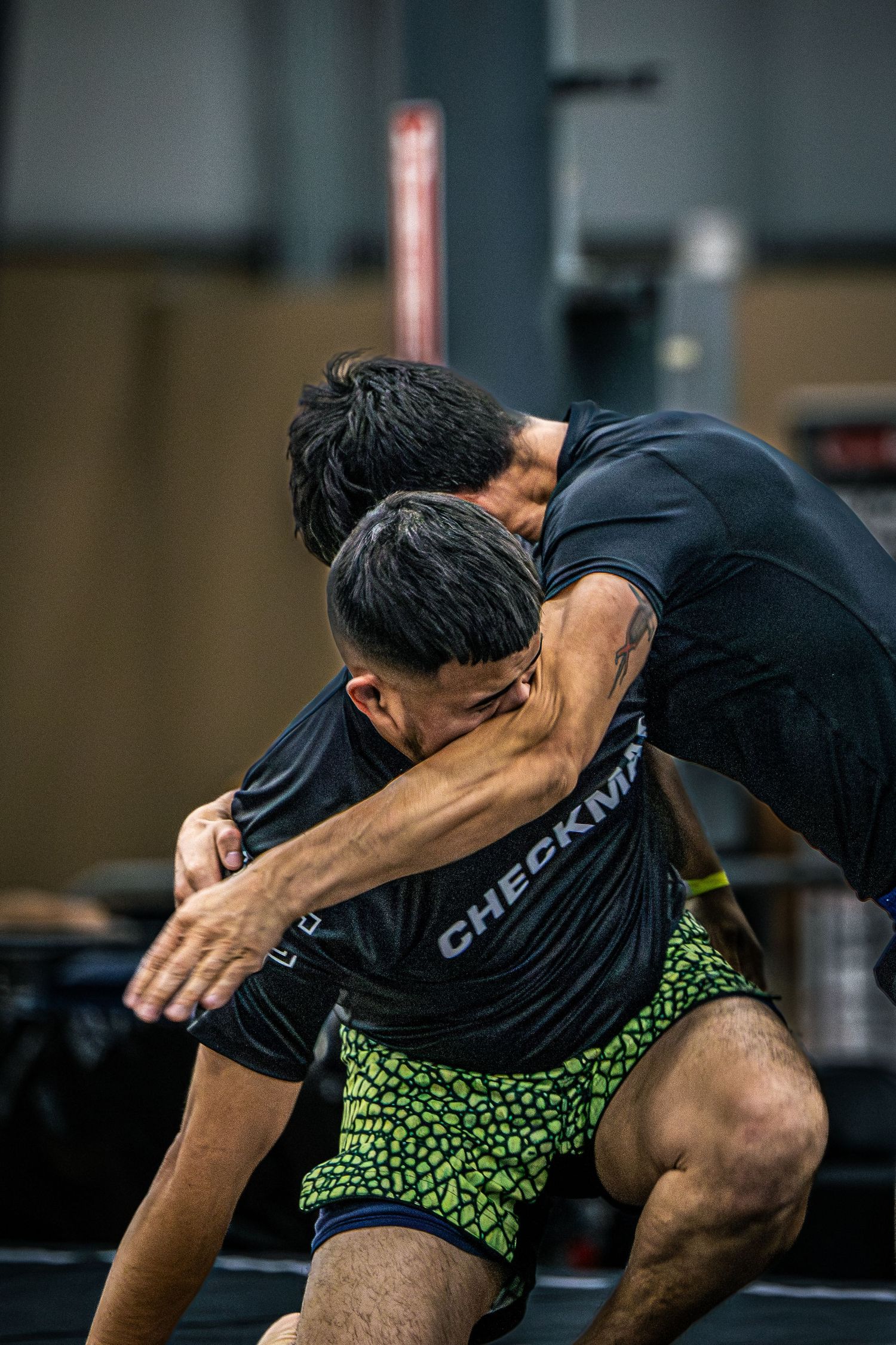 Two athletes in black Checkmat shirts practice wrestling grappling techniques in a gym.