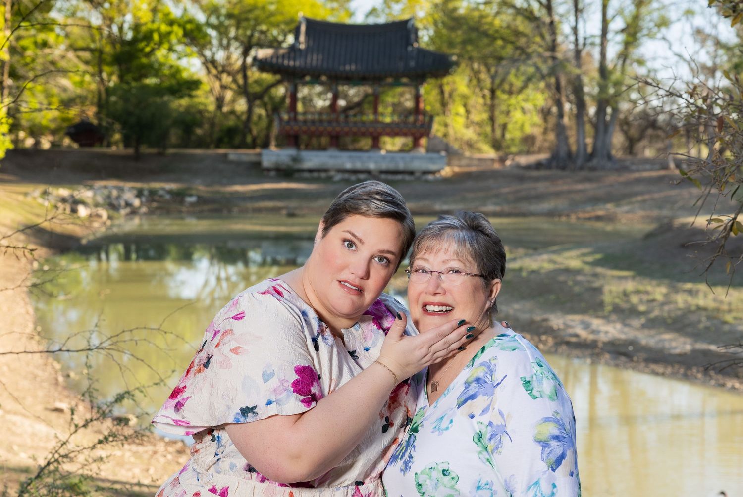Two women wearing floral dresses embrace in a scenic outdoor setting with a pond and trees in the background.