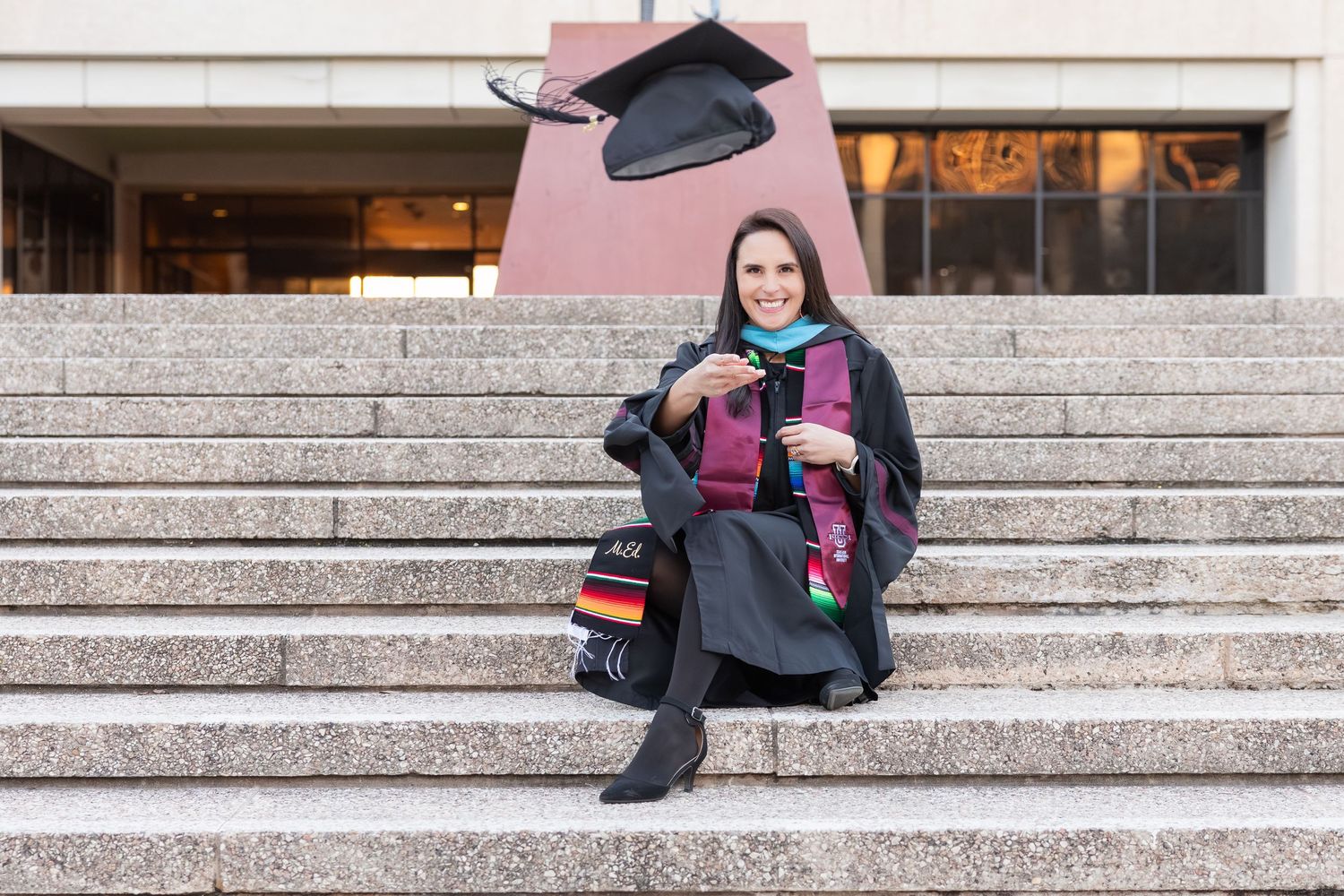 Graduate in cap and gown sits on stone steps with diploma during commencement ceremony.