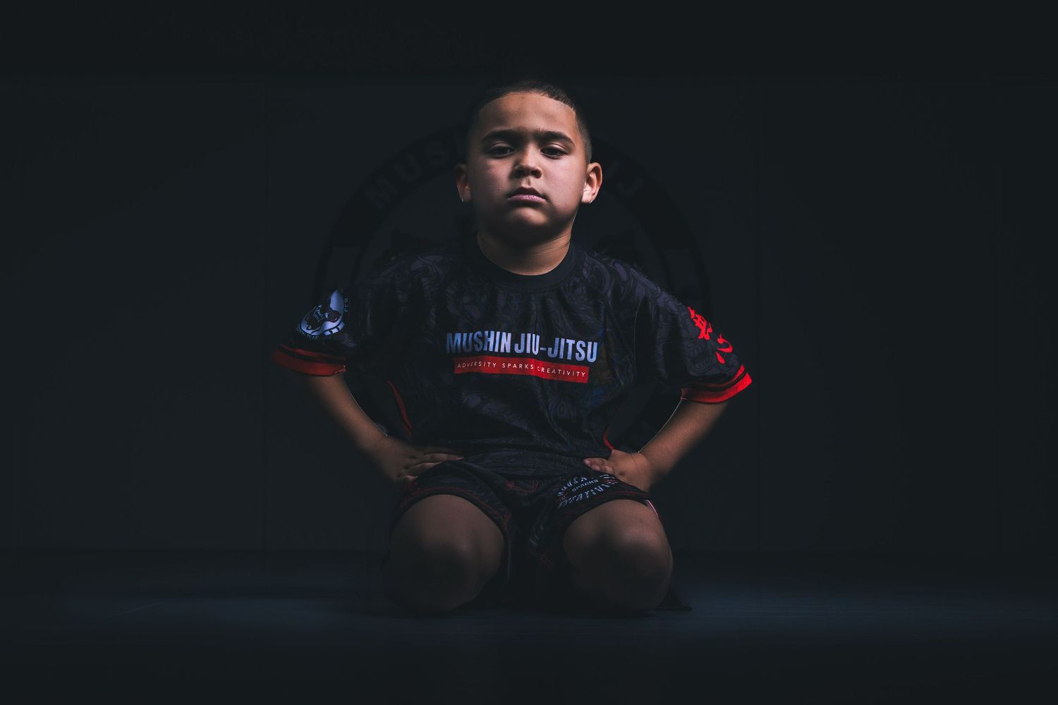 Young boy in a black Brazilian Jiu-Jitsu uniform sitting cross-legged with arms on hips against dark background.