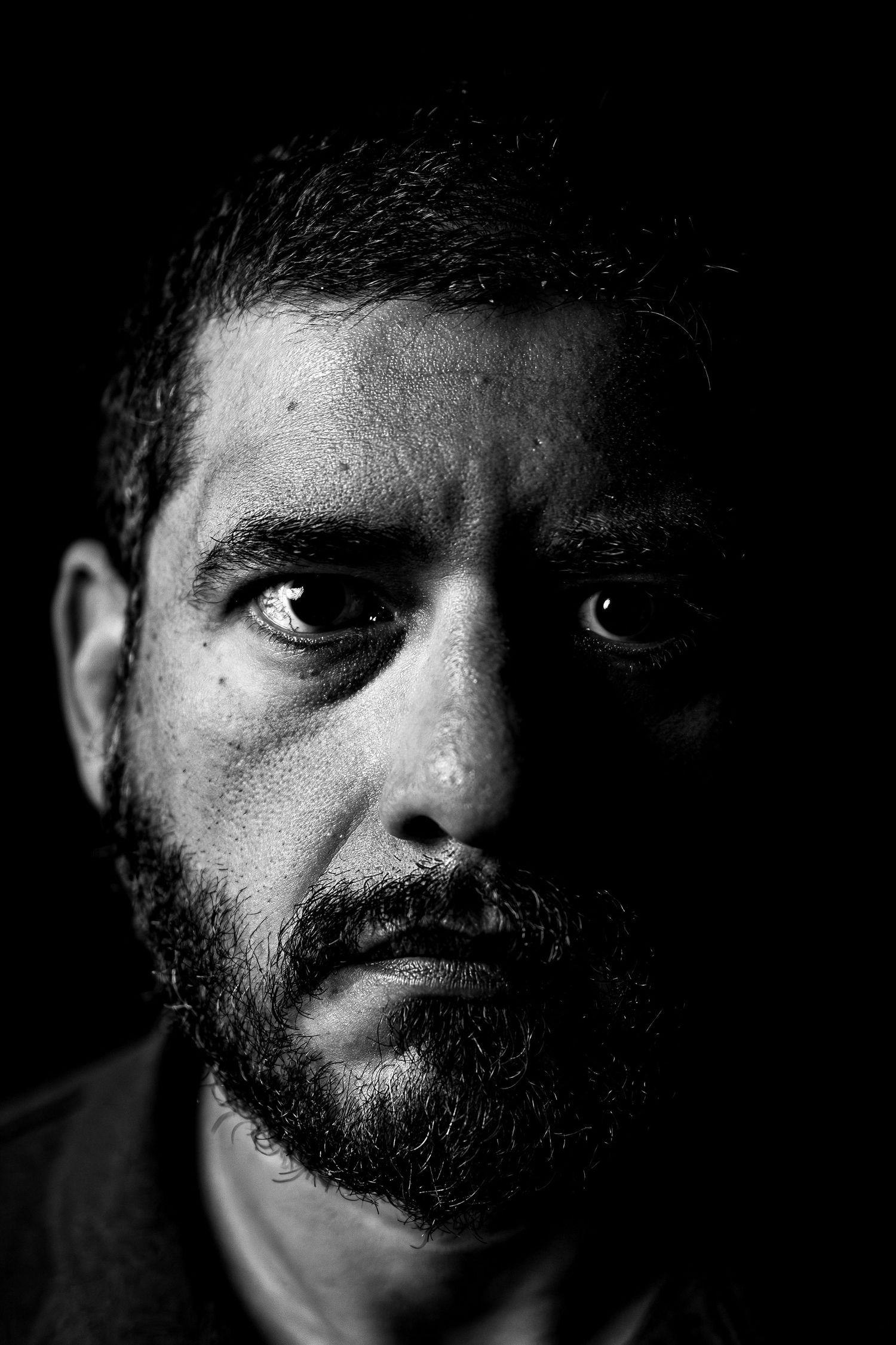 Black and white portrait of a bearded man with a tear on his cheek, conveying deep emotion against dark background.