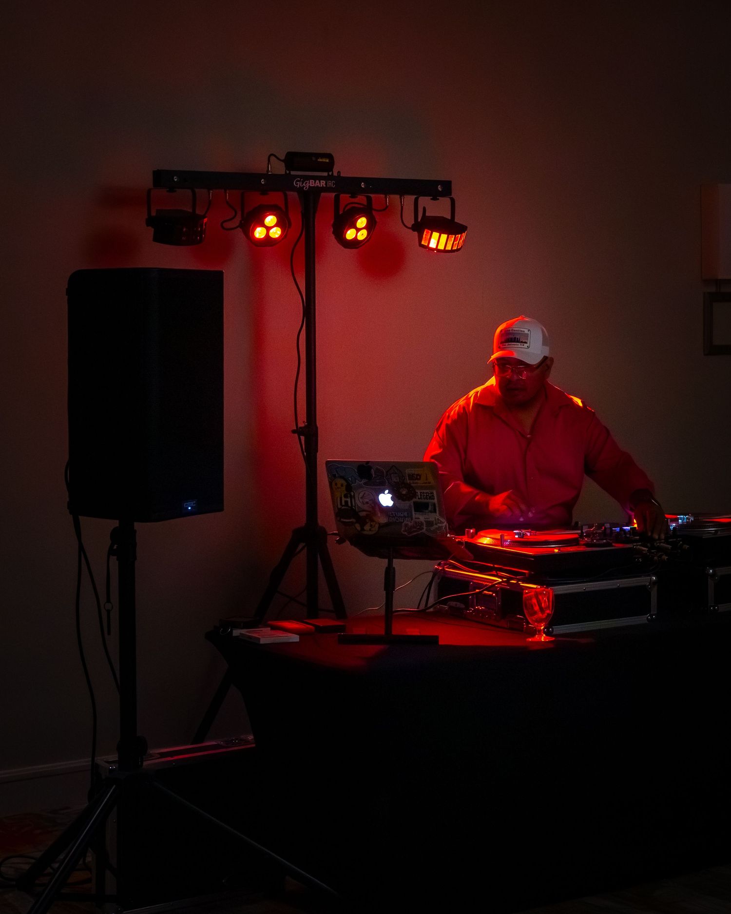 DJ performing under red stage lights with speaker, lighting rig, turntables, and laptop in a dimly lit venue.
