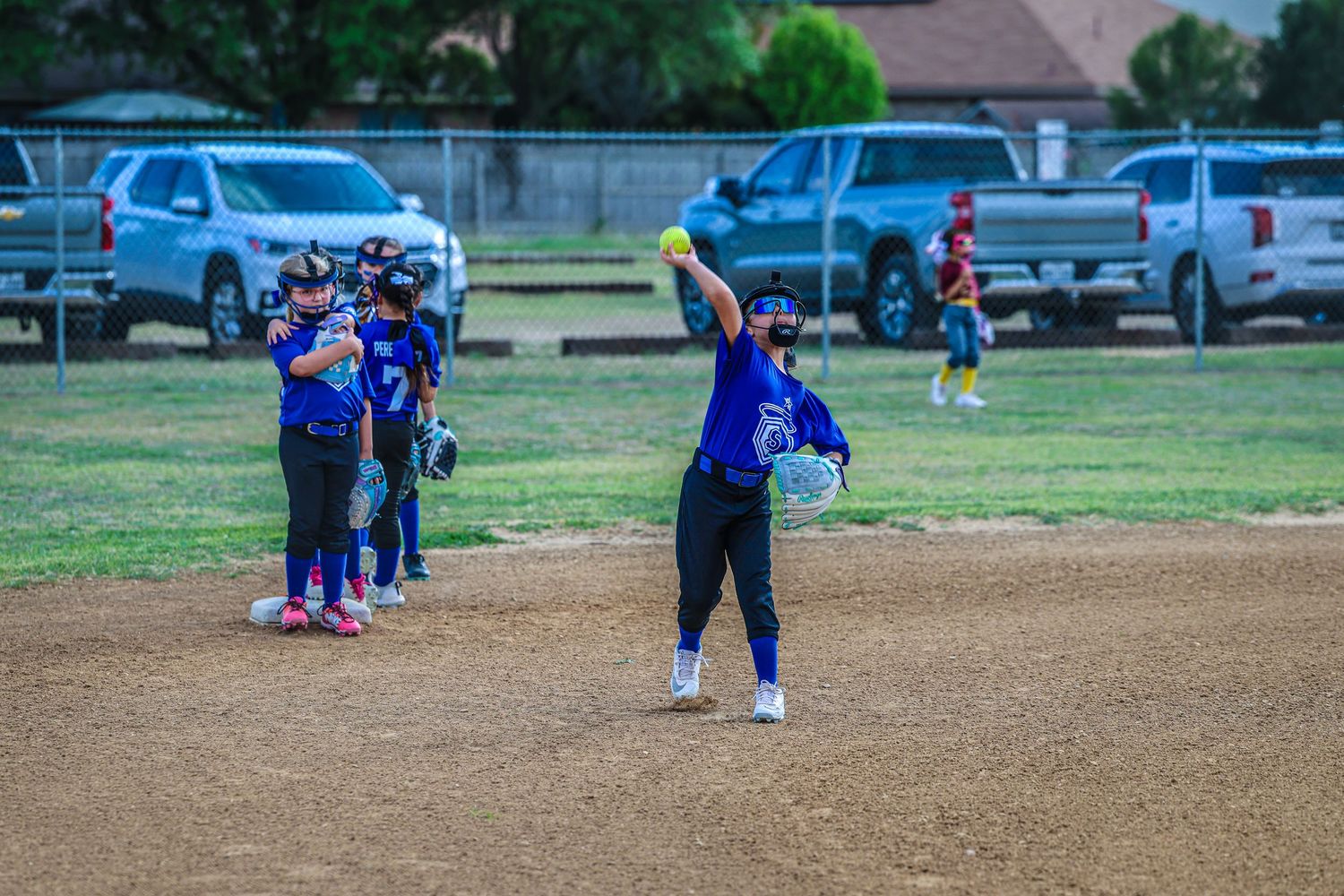 Youth softball pitcher winds up to throw during a game with teammates gathered on the field behind her.