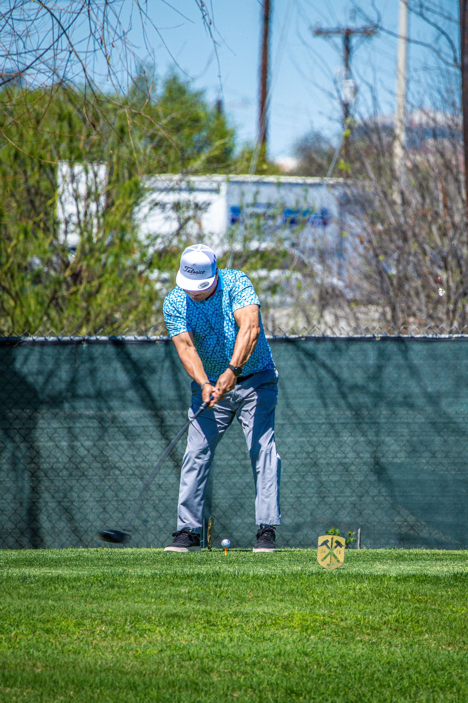 Golfer in blue shirt preparing to swing on a green course near a fence on a sunny day.