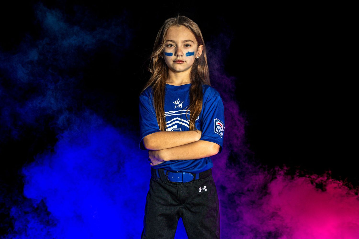 Young girl in blue softball uniform with face paint stands confidently with arms crossed against colorful smoke background.