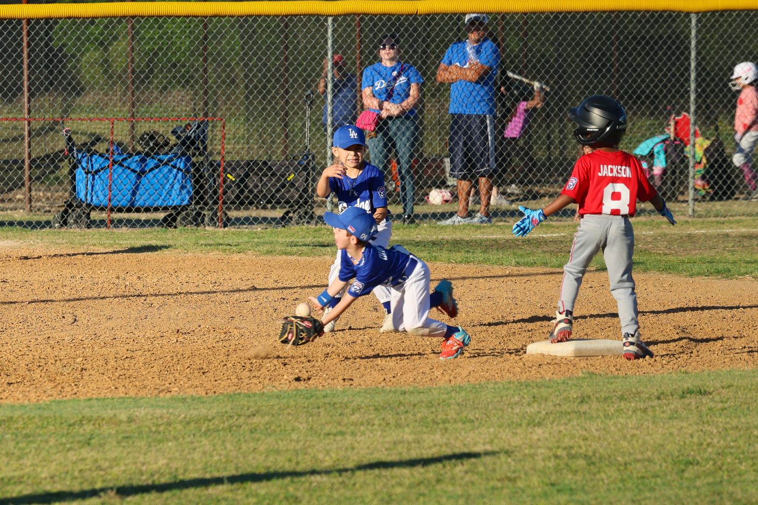 Youth baseball player in blue uniform fielding at base as red team runner approaches during a little league game.