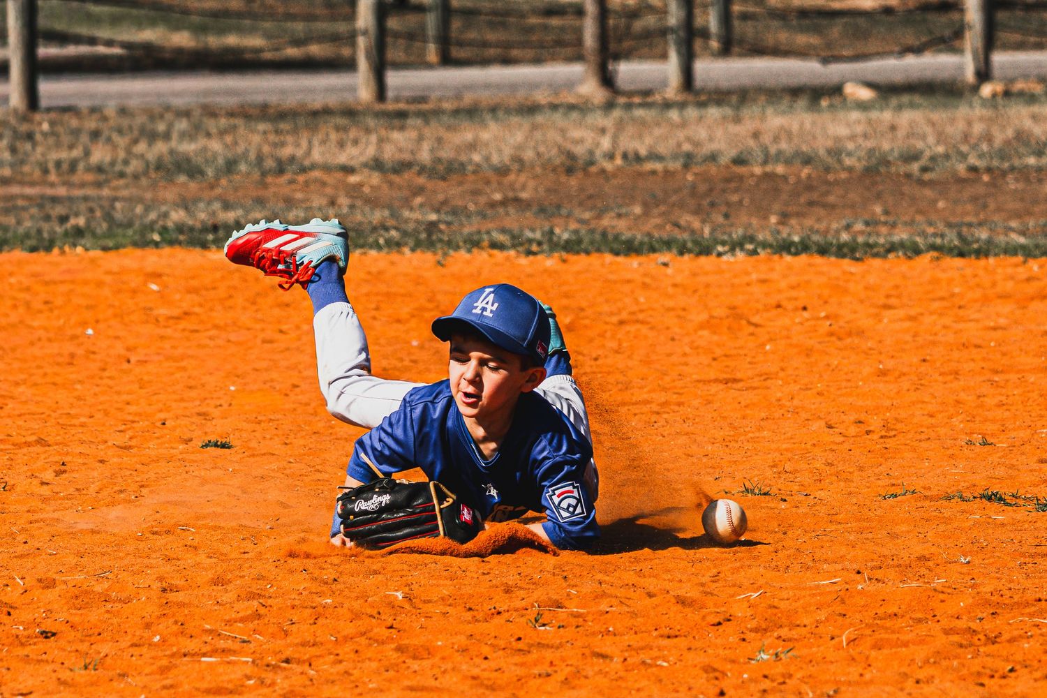 Young baseball player diving on orange dirt field attempting to field a ground ball while wearing blue Dodgers uniform.