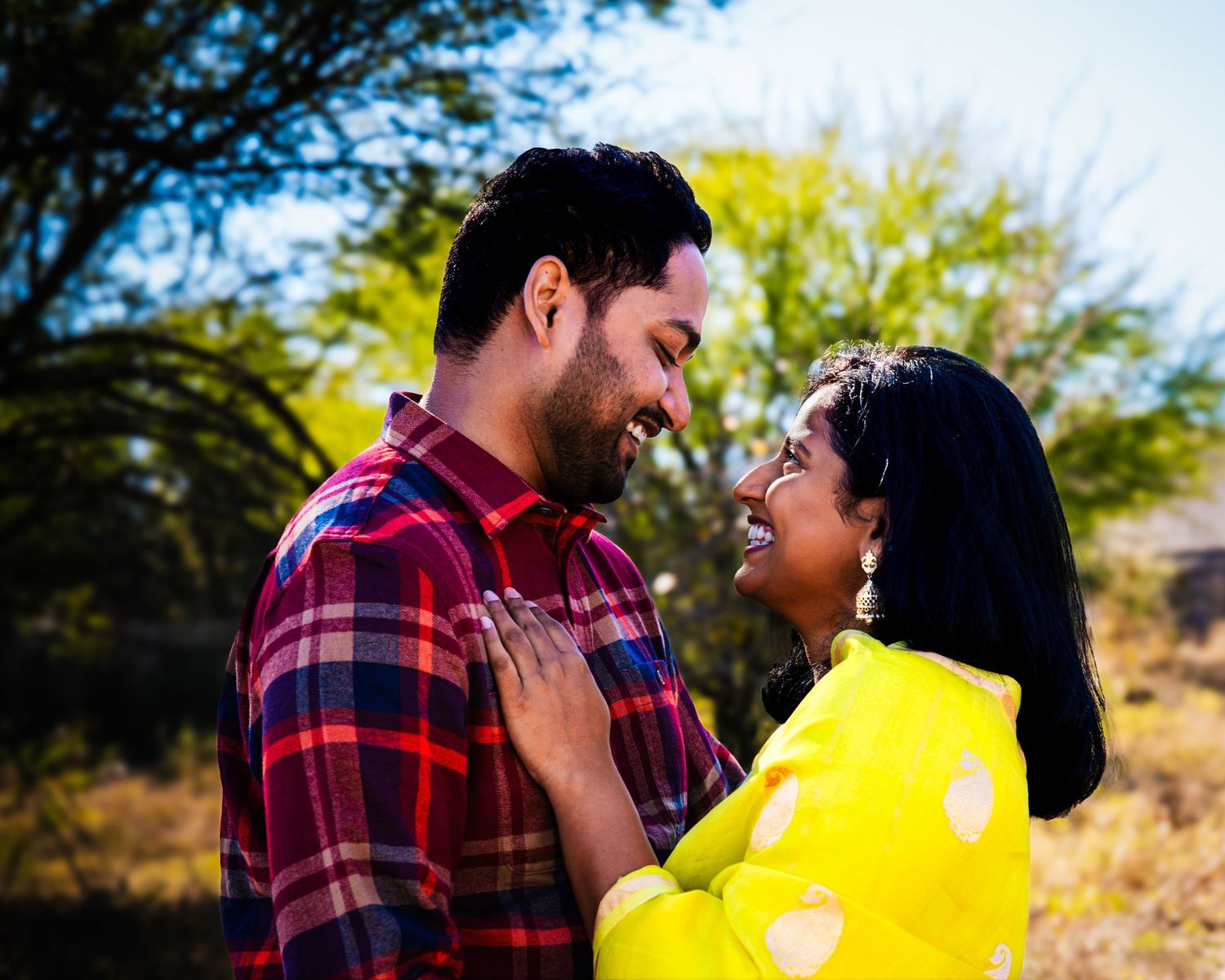 A couple in a plaid shirt and yellow dress share a romantic moment outdoors during golden hour.
