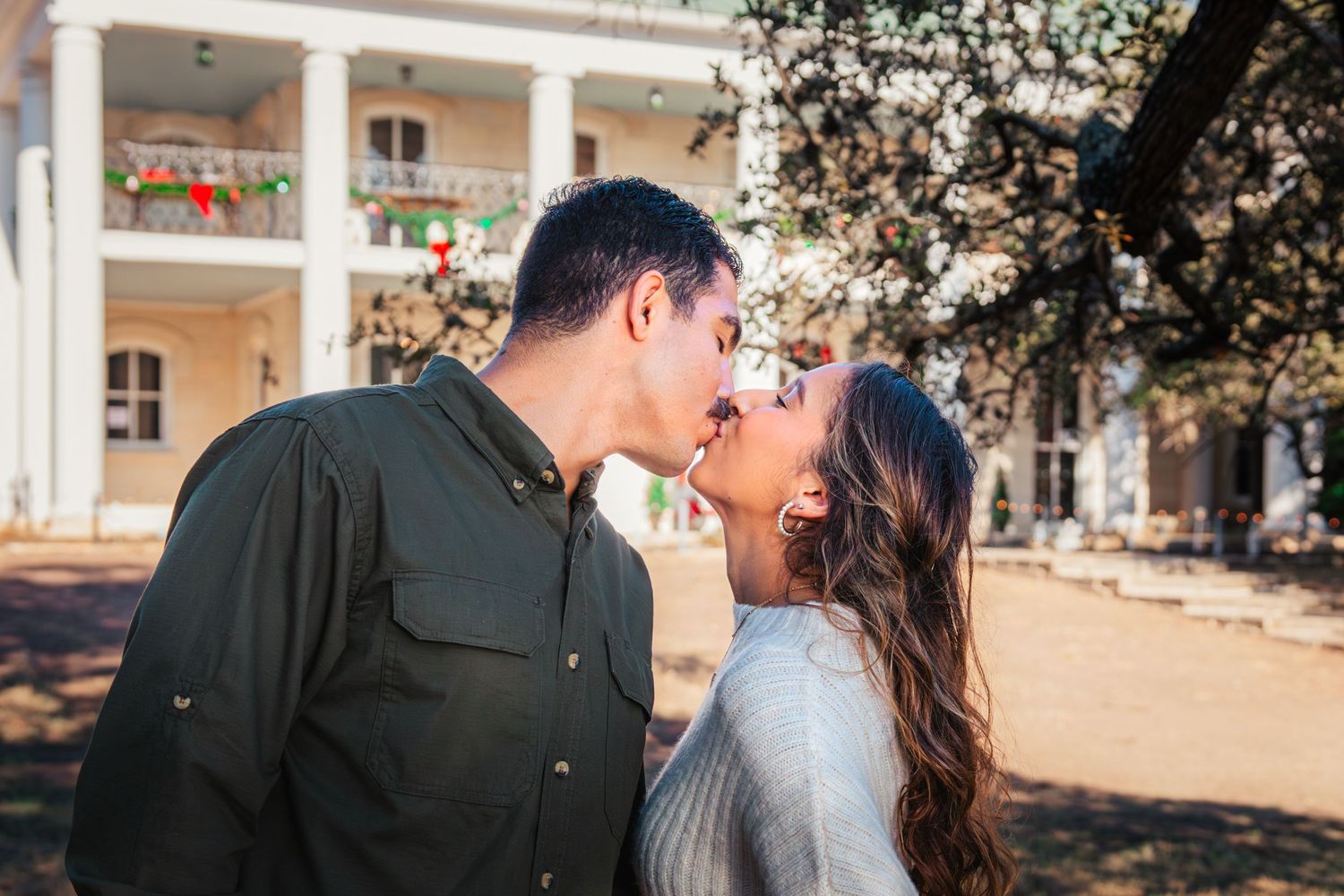 A romantic couple shares a tender kiss on the grounds of a stately southern mansion with white columns.