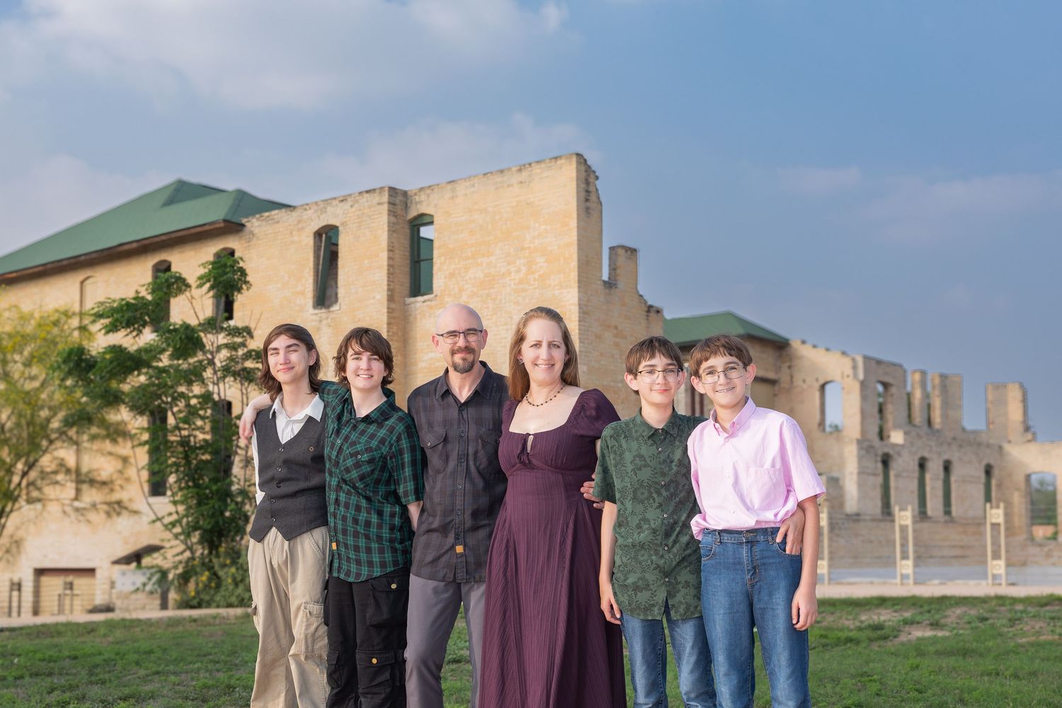 Family of six posing outdoors in front of historic stone ruins on a green lawn under a cloudy sky.