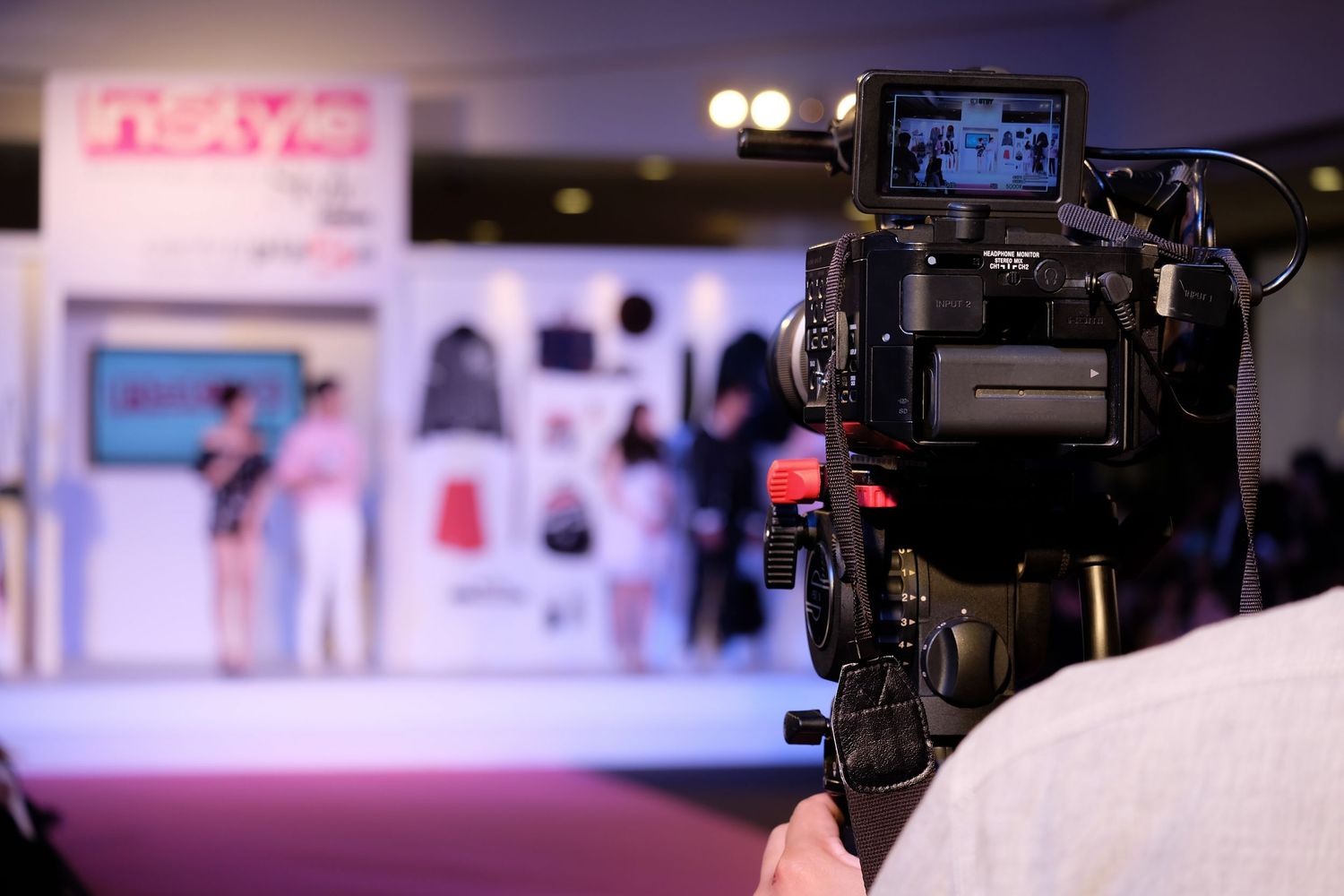 Professional video camera filming a fashion show runway event with colorful clothing displays on stage.