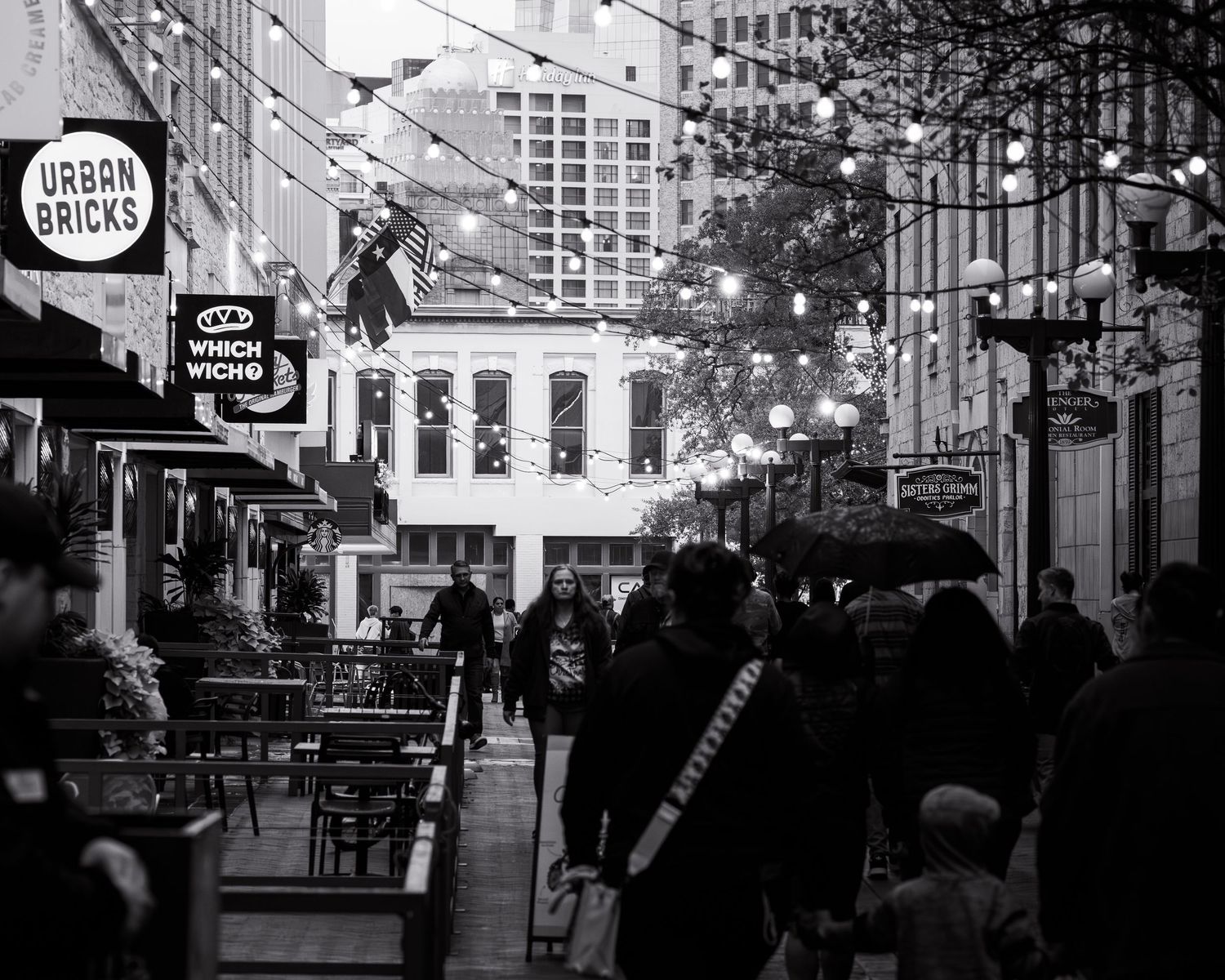 Black and white urban street scene with pedestrians walking past storefronts and traffic signals.