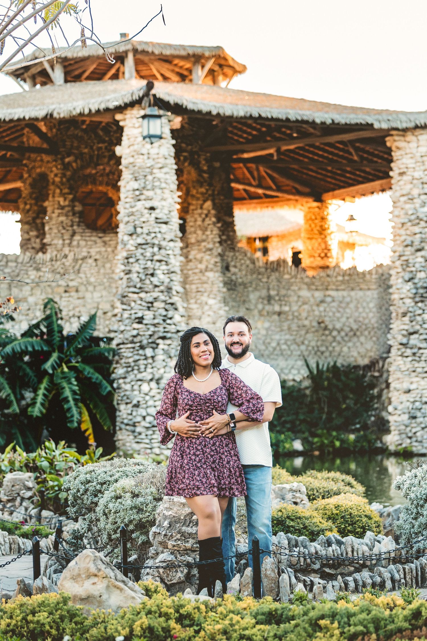 A couple poses together in front of rustic stone walls and lush garden landscaping at sunset.