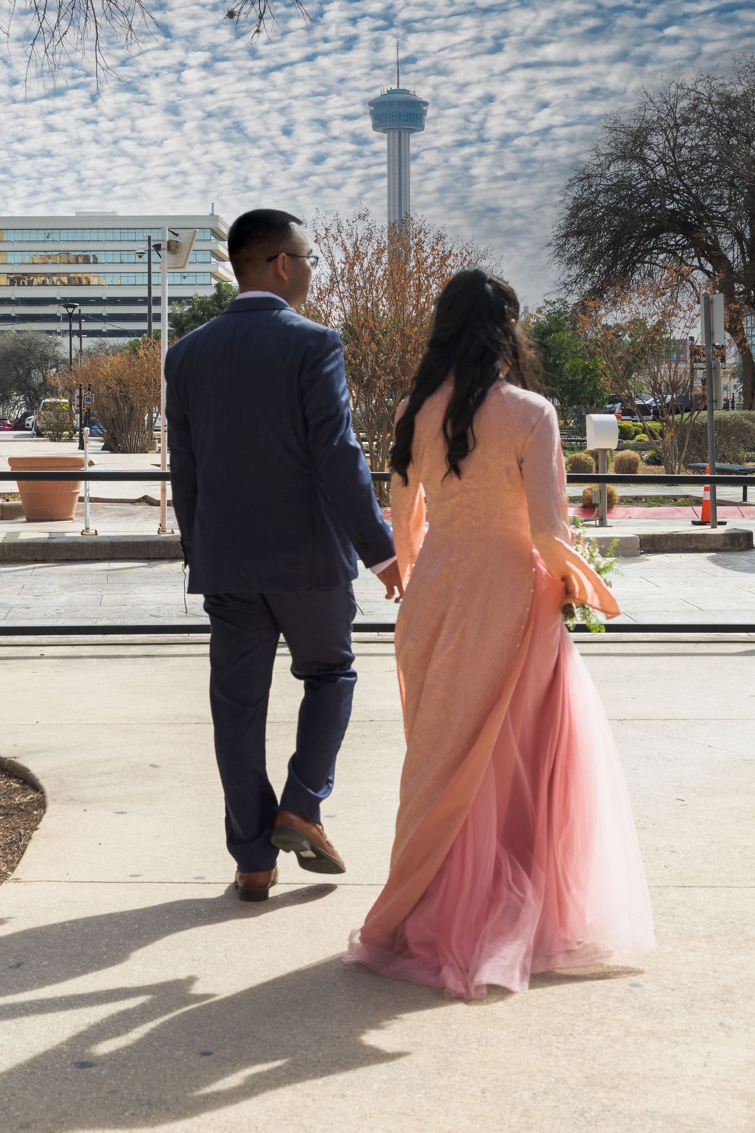 A couple in formal attire walk together on a path with one wearing a navy suit and the other in a flowing pink gown.