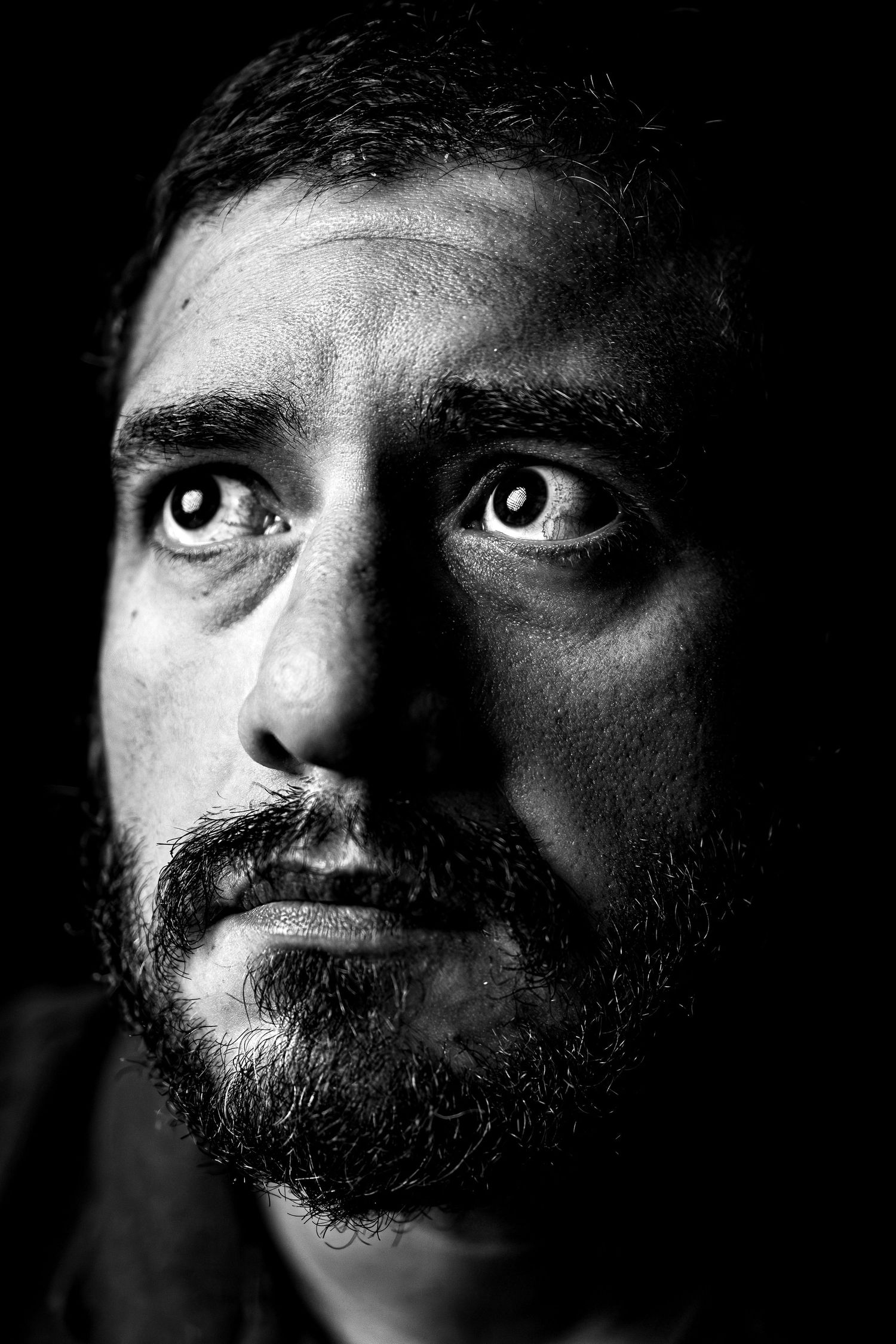 Black and white portrait of a bearded man with intense wide eyes looking up against a dark background.