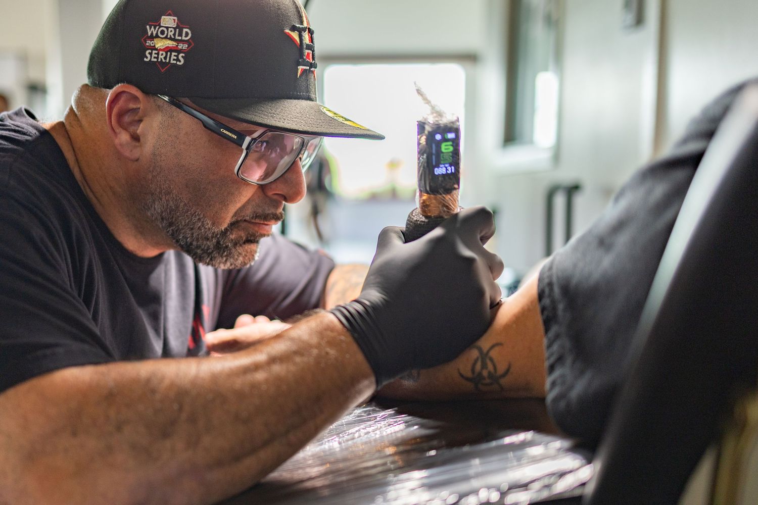 A tattoo artist wearing black gloves and an Astros cap carefully works on a detailed tattoo design on a client.
