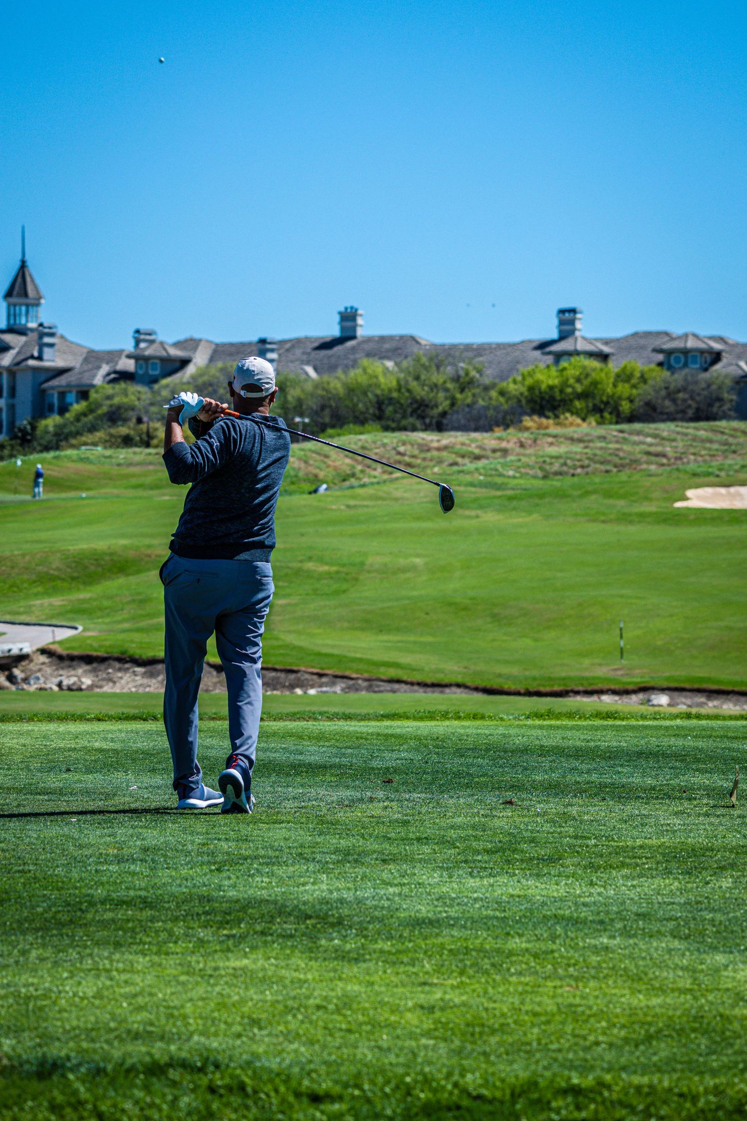 Golfer mid-swing on a lush green golf course with sand bunkers and clubhouse buildings under a clear blue sky.
