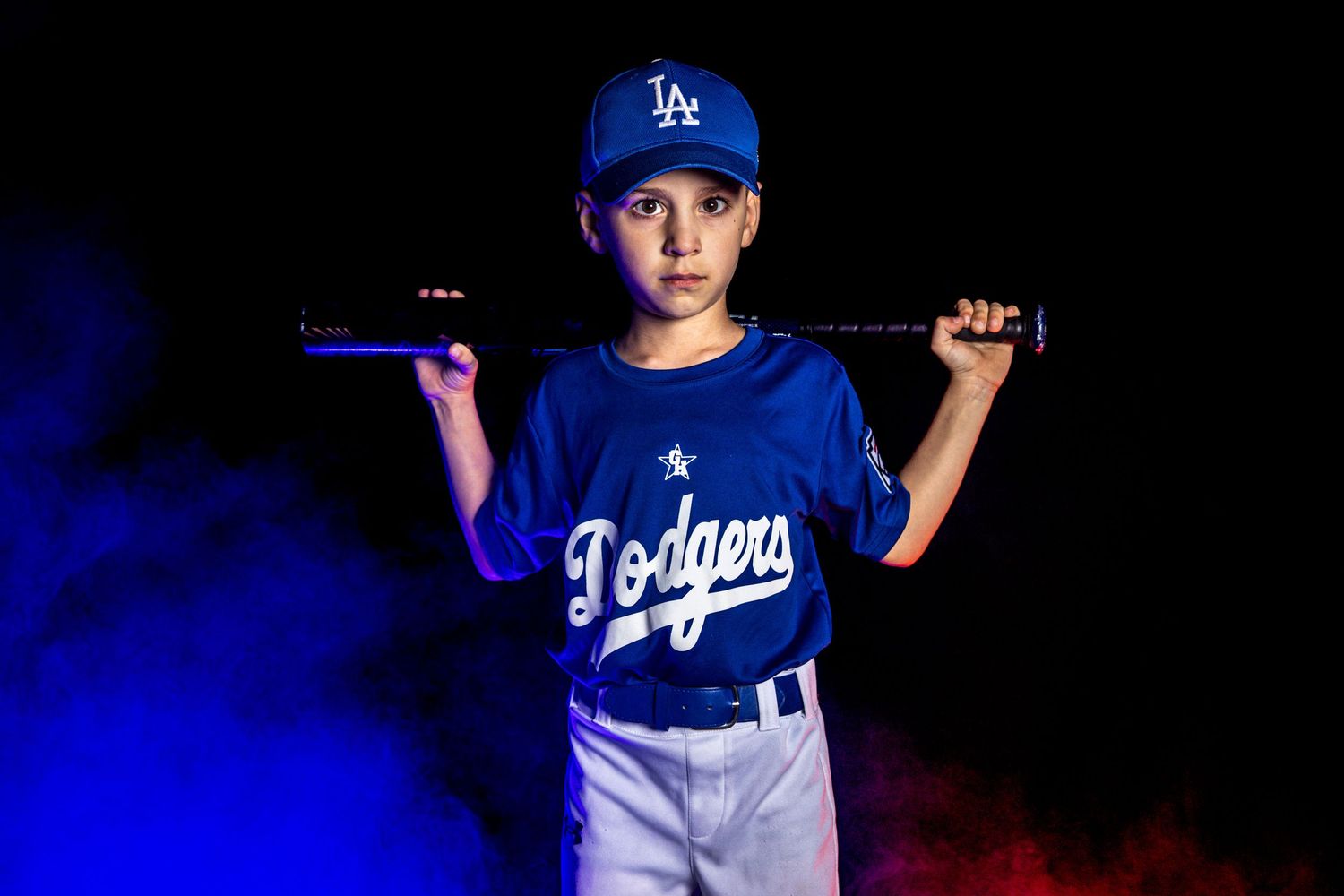 Young boy in LA Dodgers uniform holding baseball bat on shoulders against dramatic dark background.