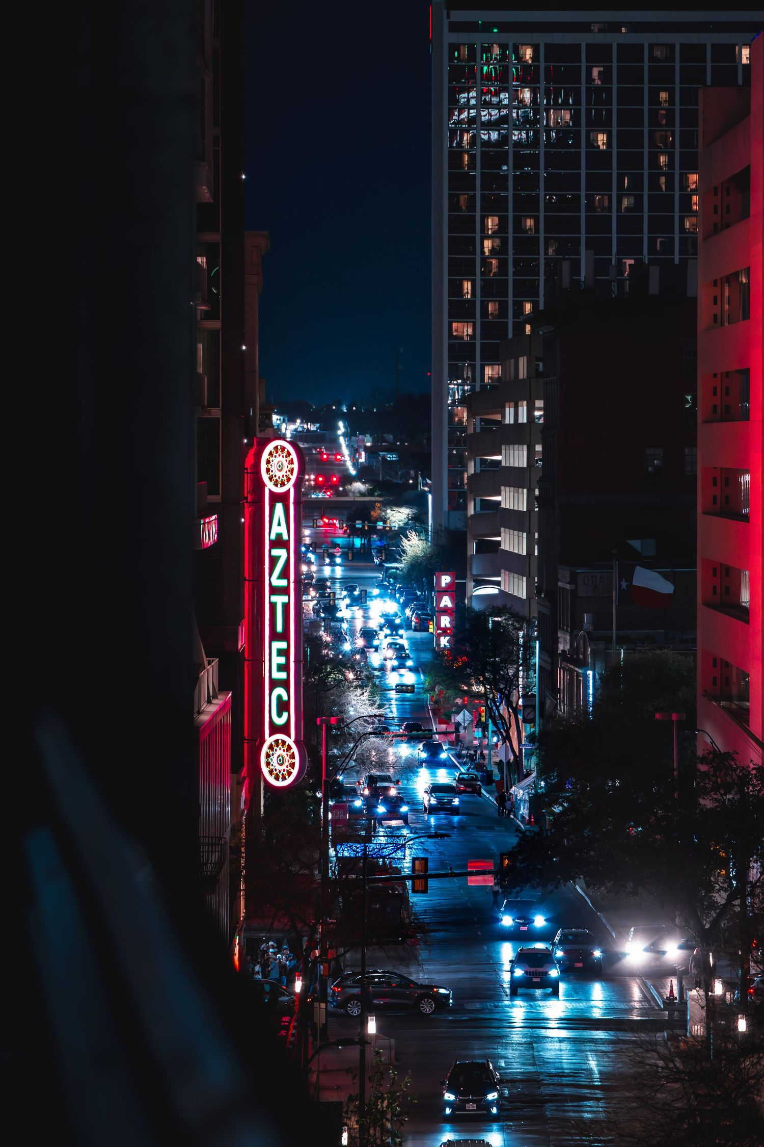 Nighttime cityscape with glowing Aztec Theater neon sign illuminating a busy downtown street lined with cars and skyscrapers.