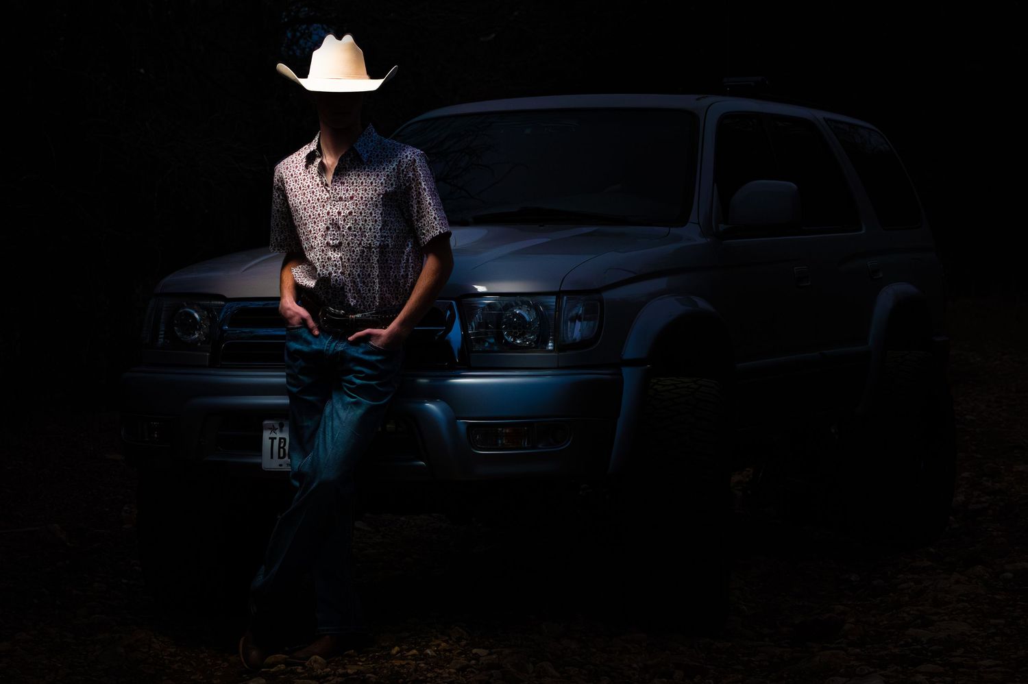 Man in cowboy hat leaning on silver SUV truck at night in dark outdoor setting.