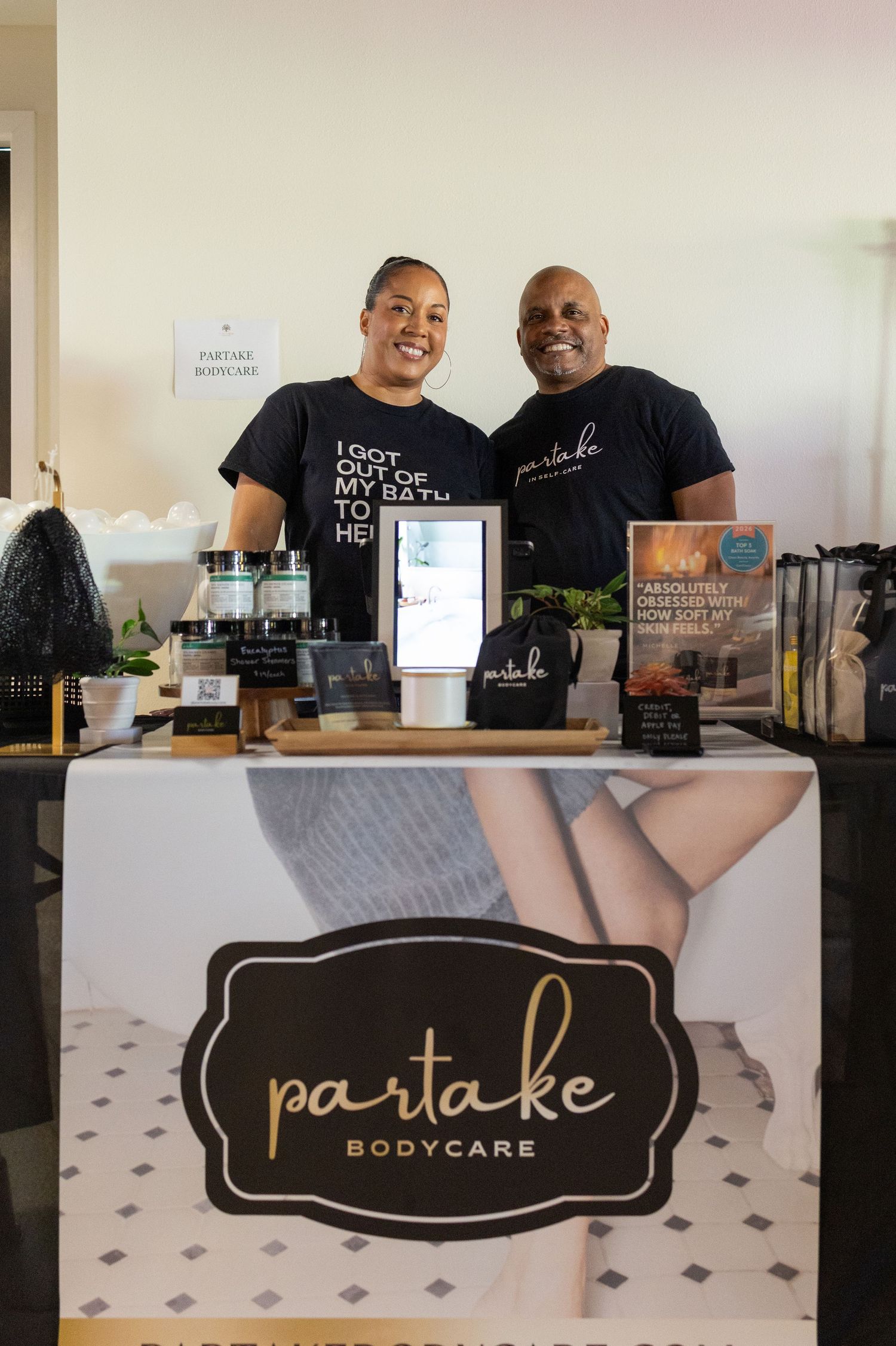 Two smiling owners stand behind a Partake Bodycare branded booth displaying skincare products at a vendor event.