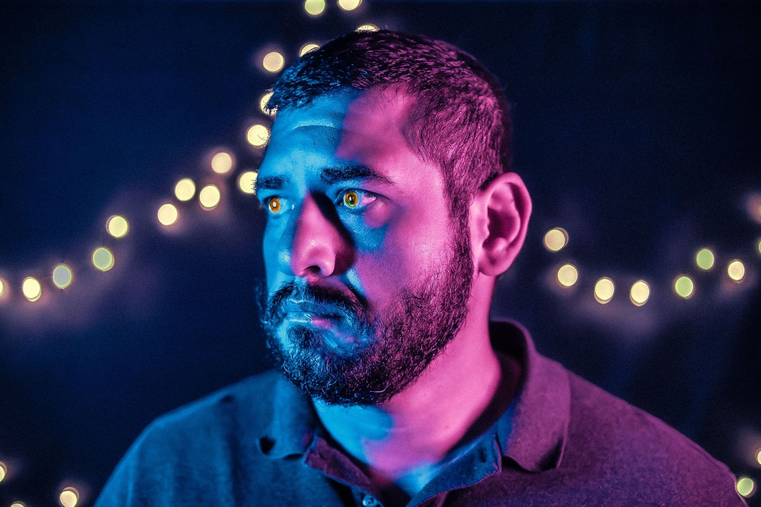 Man with beard under blue and purple neon lights with bokeh string lights in background.