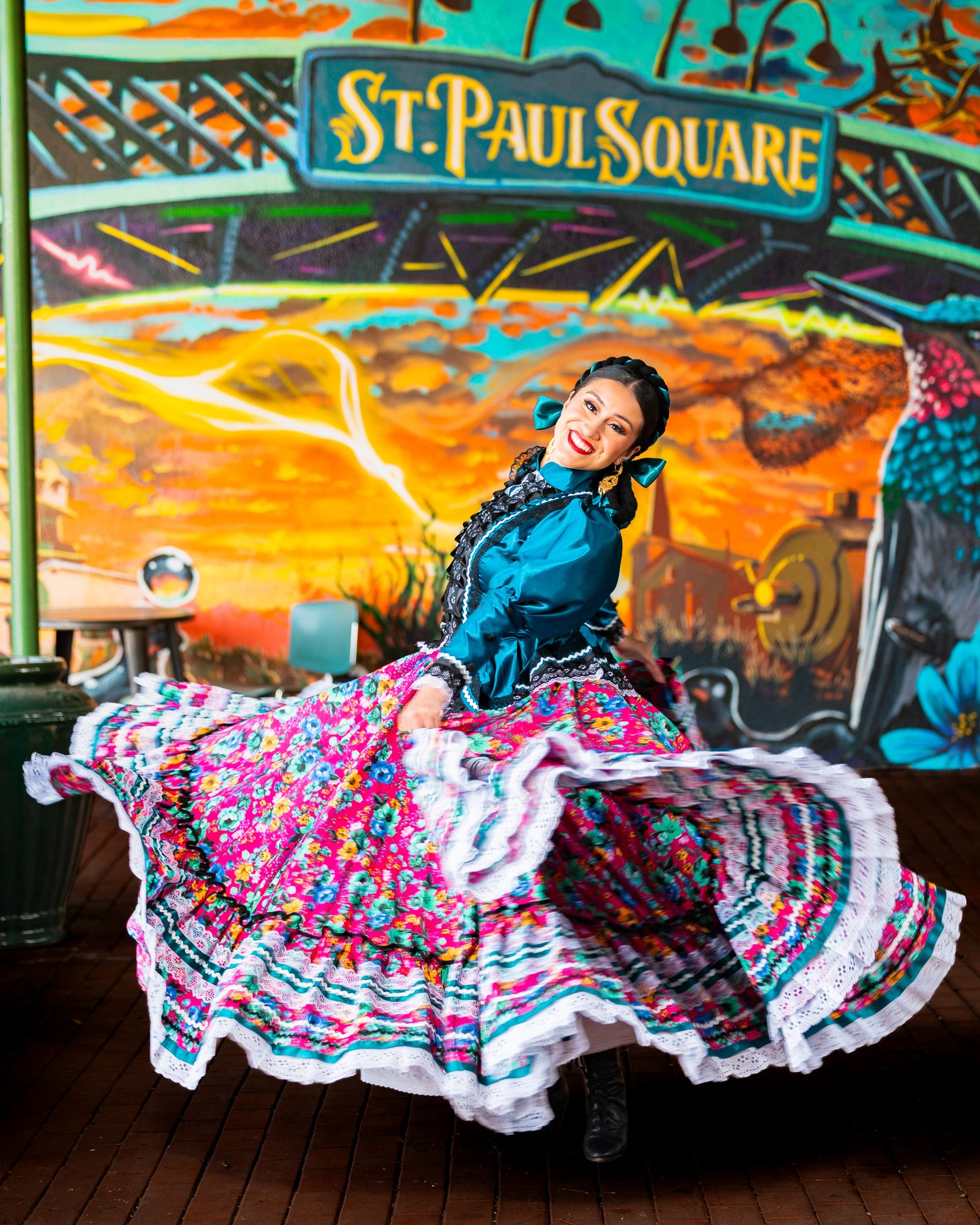 Dancer in vibrant traditional Mexican dress twirls under St. Paul Square sign with colorful backdrop at sunset.