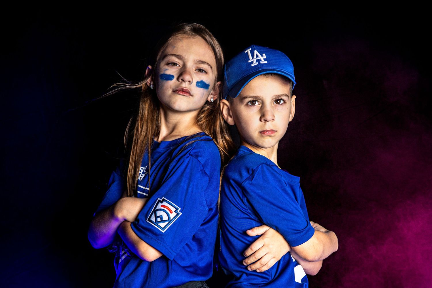 Two kids in LA Dodgers uniforms posing back-to-back with blue face paint in dramatic lighting.