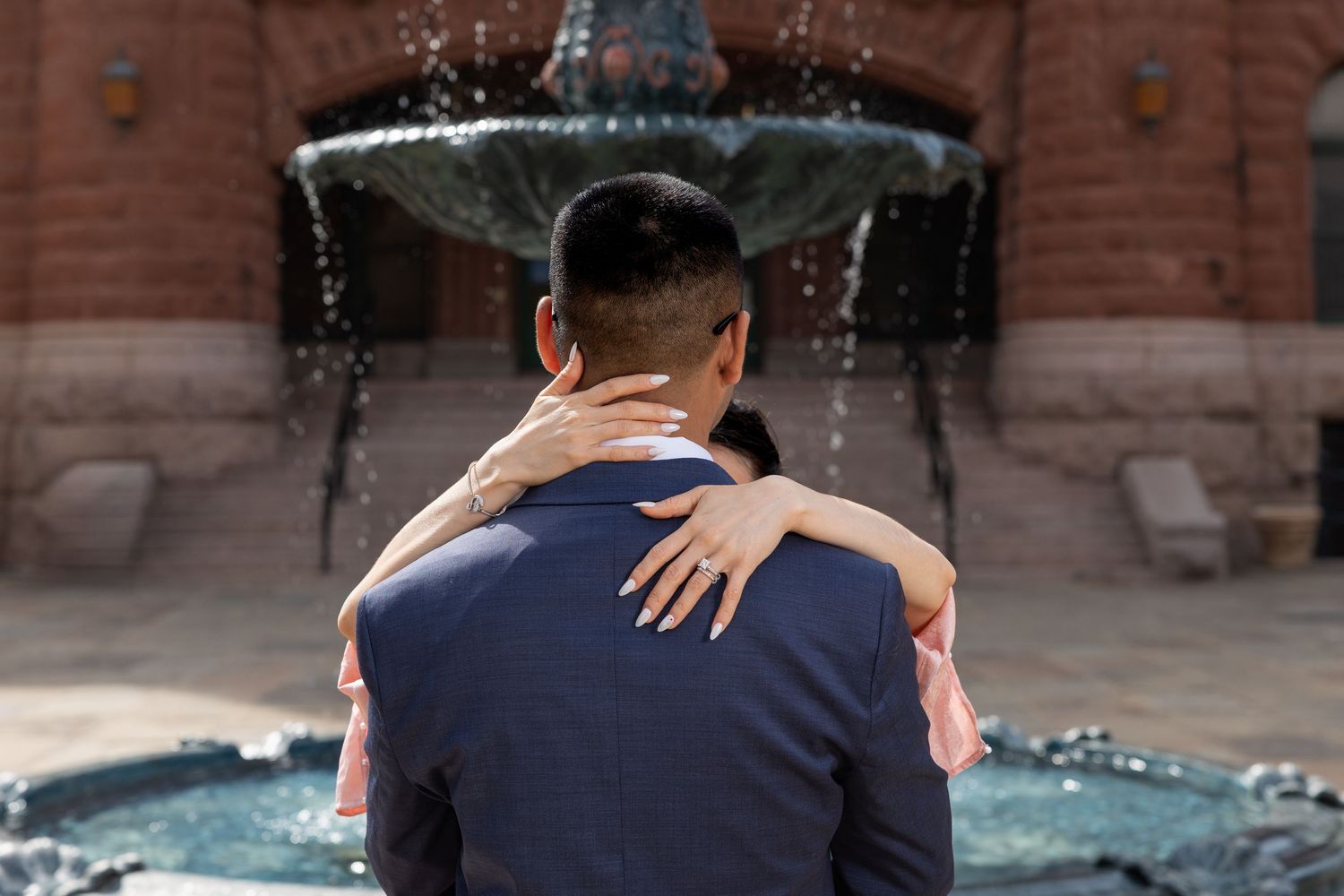 A couple embraces while dancing in front of a fountain in an urban plaza at sunset.