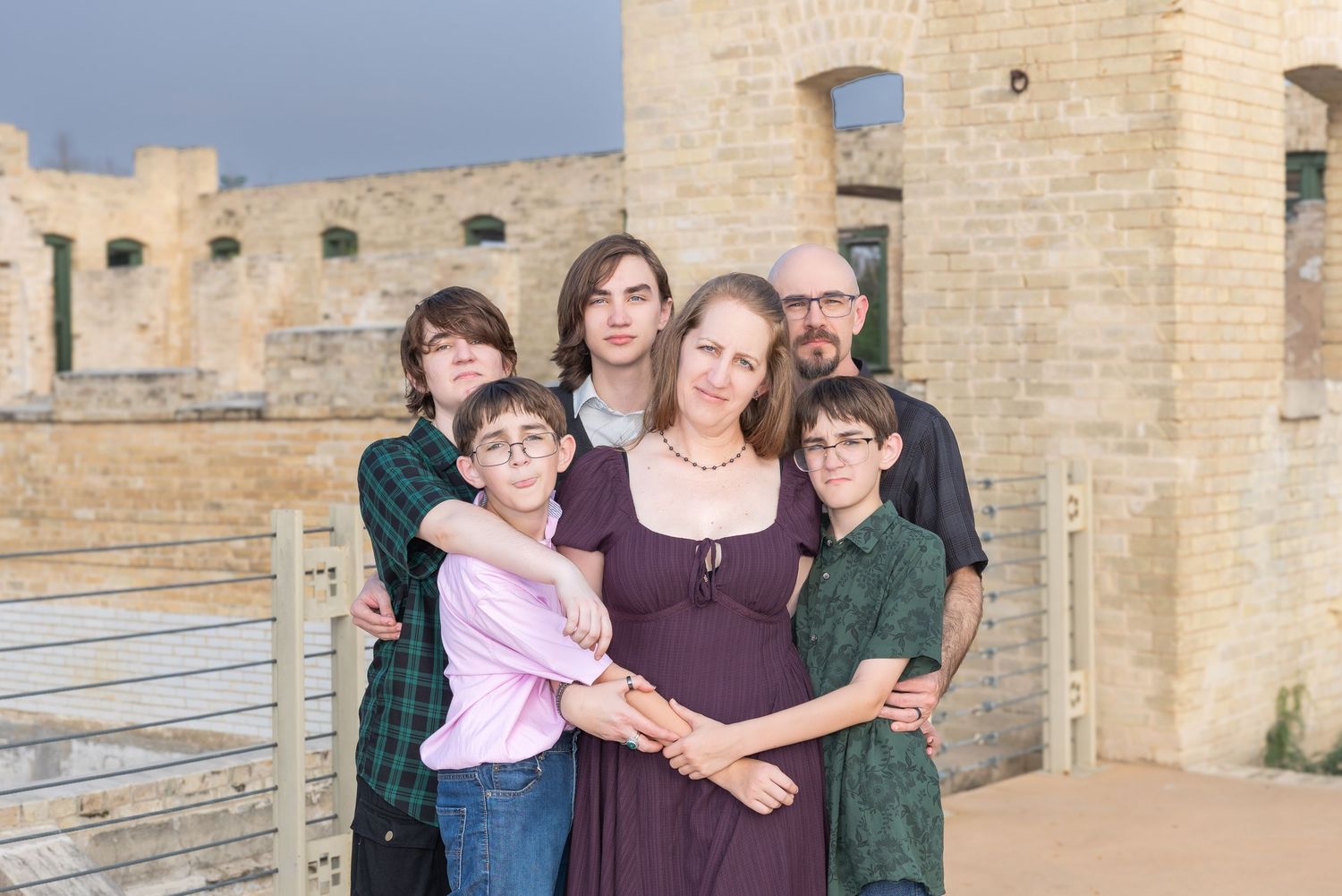 Family of six posing together outdoors near a stone building under a cloudy sky.