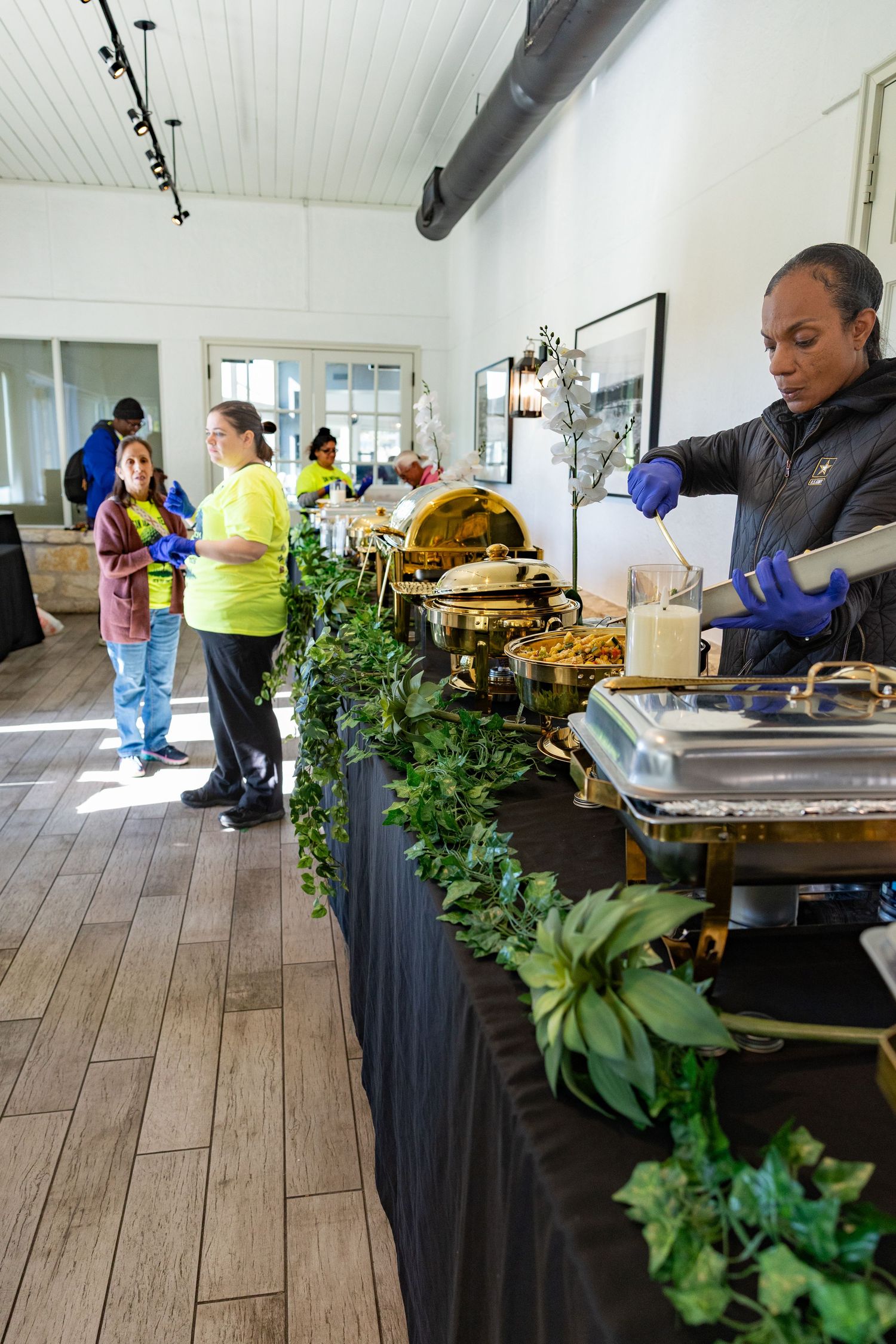 Catering staff serving food at a buffet line with chafing dishes and green garland in an elegant event venue.