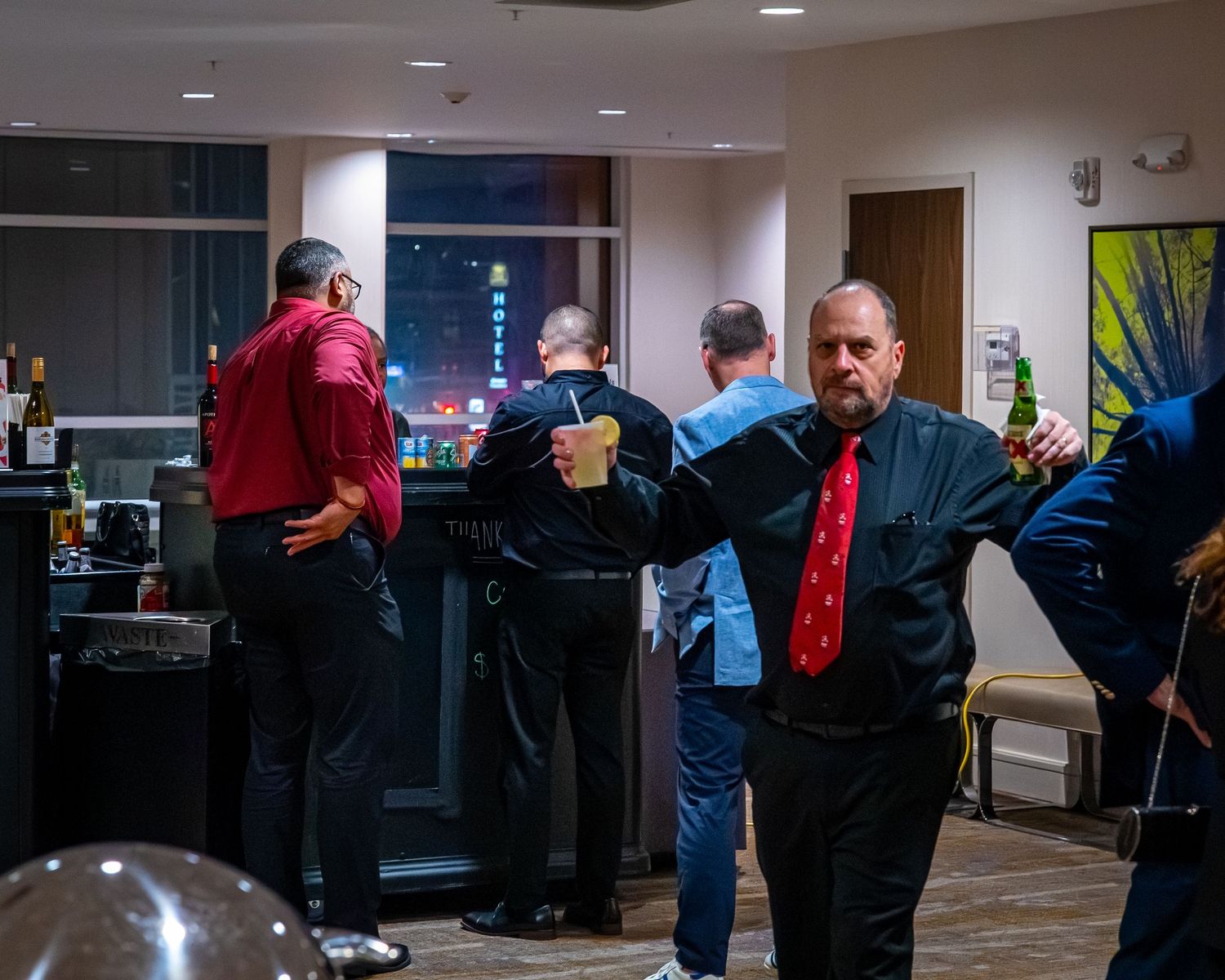 Men in formal attire socializing at an indoor evening party with city lights visible through windows.