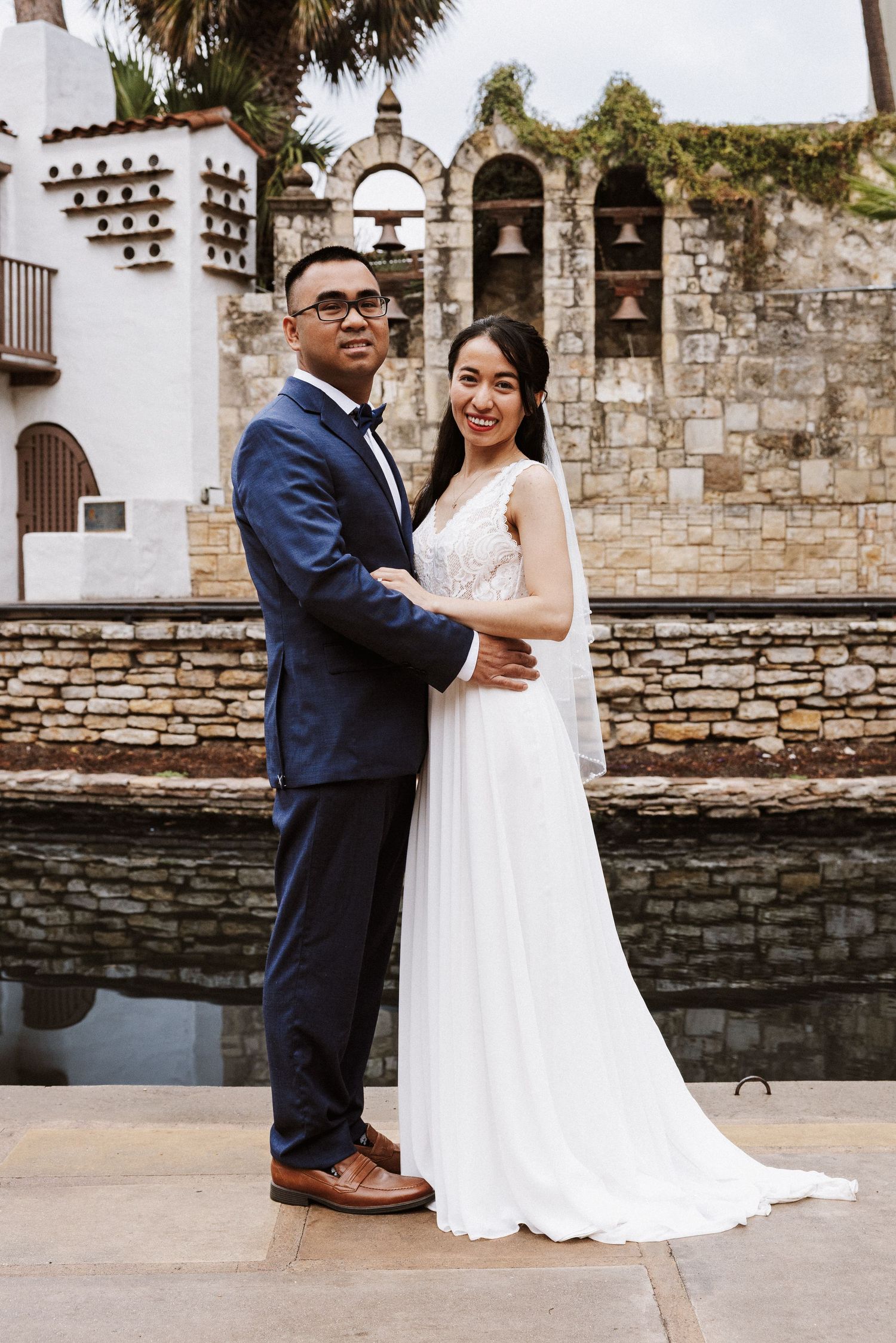 A couple in wedding attire poses together against a rustic stone wall backdrop at a historic venue.