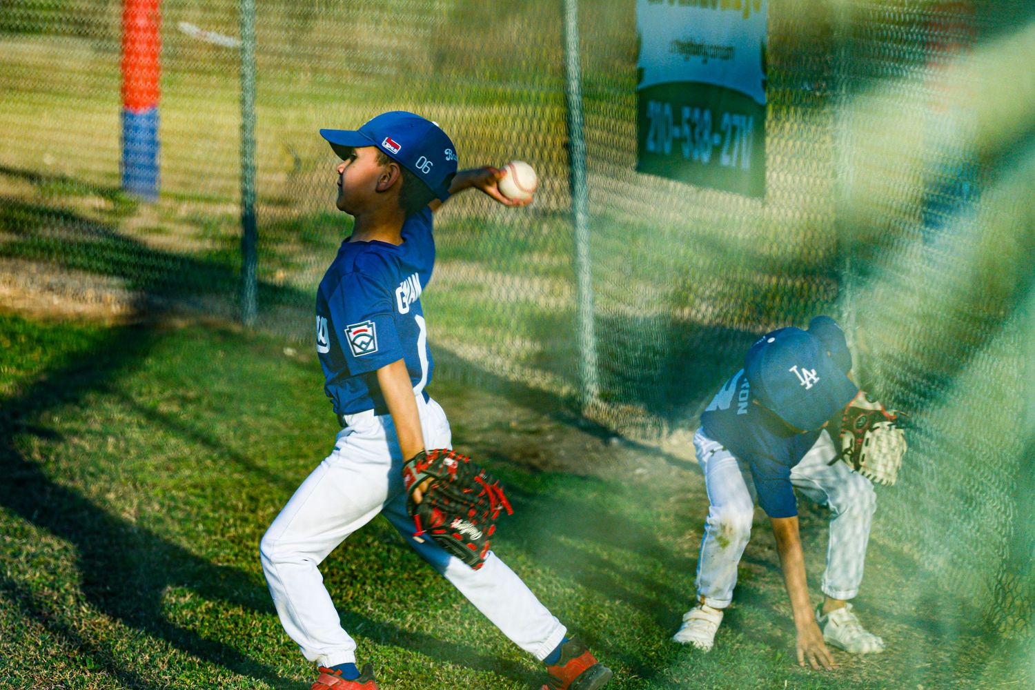 Young baseball players in blue uniforms practicing pitching and fielding behind batting cage net.