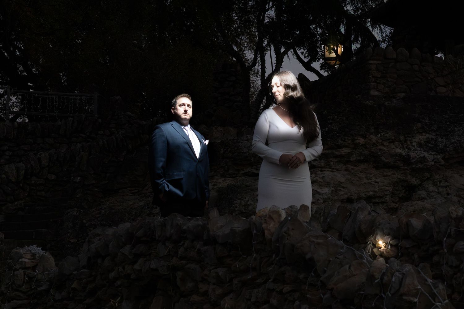 A bride and groom stand illuminated in a dark forest setting, dressed in white and navy blue wedding attire.